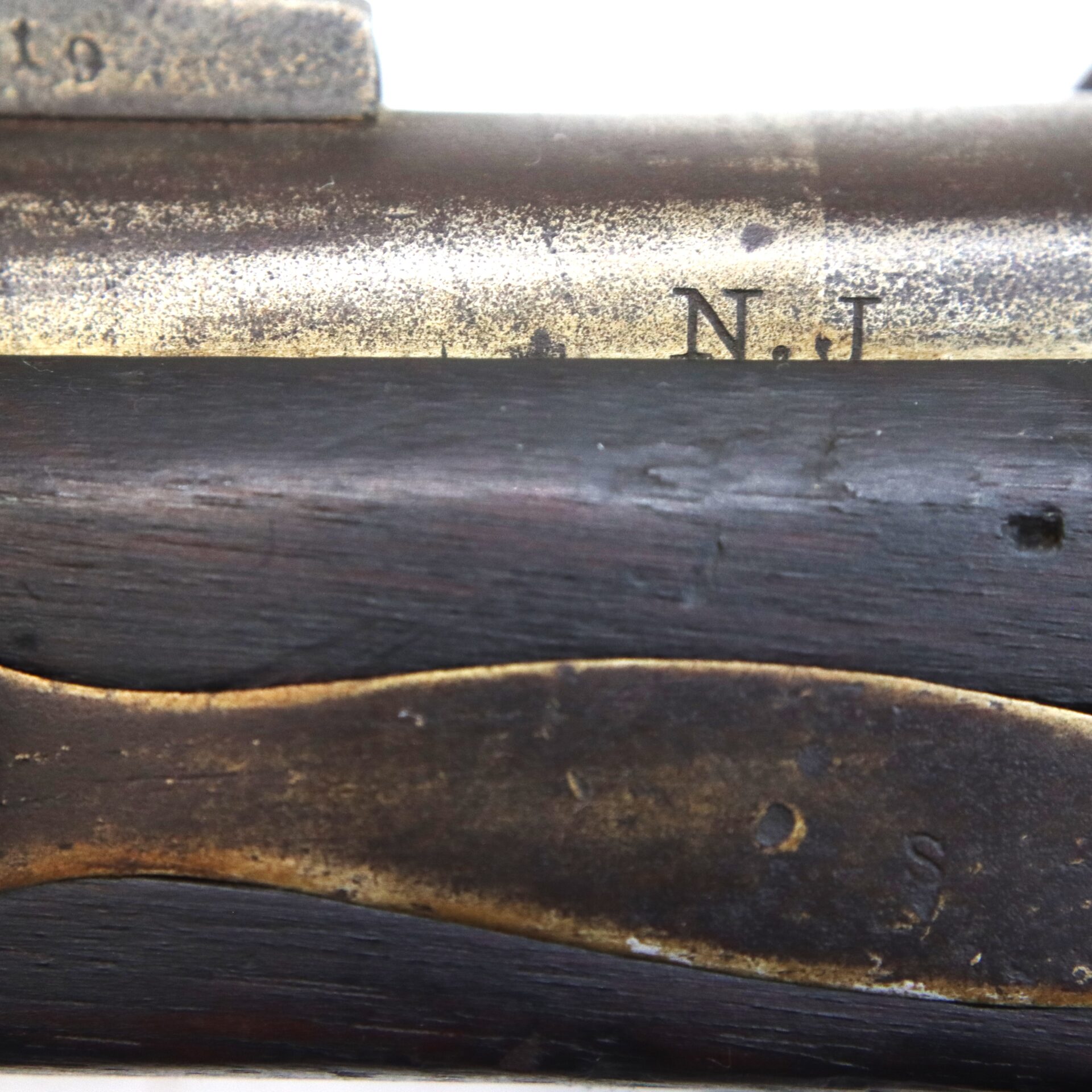 Close-up of an aged firearm showing part of the metal receiver marked with the letters "N.J." above a dark wooden stock with brass or bronze hardware.