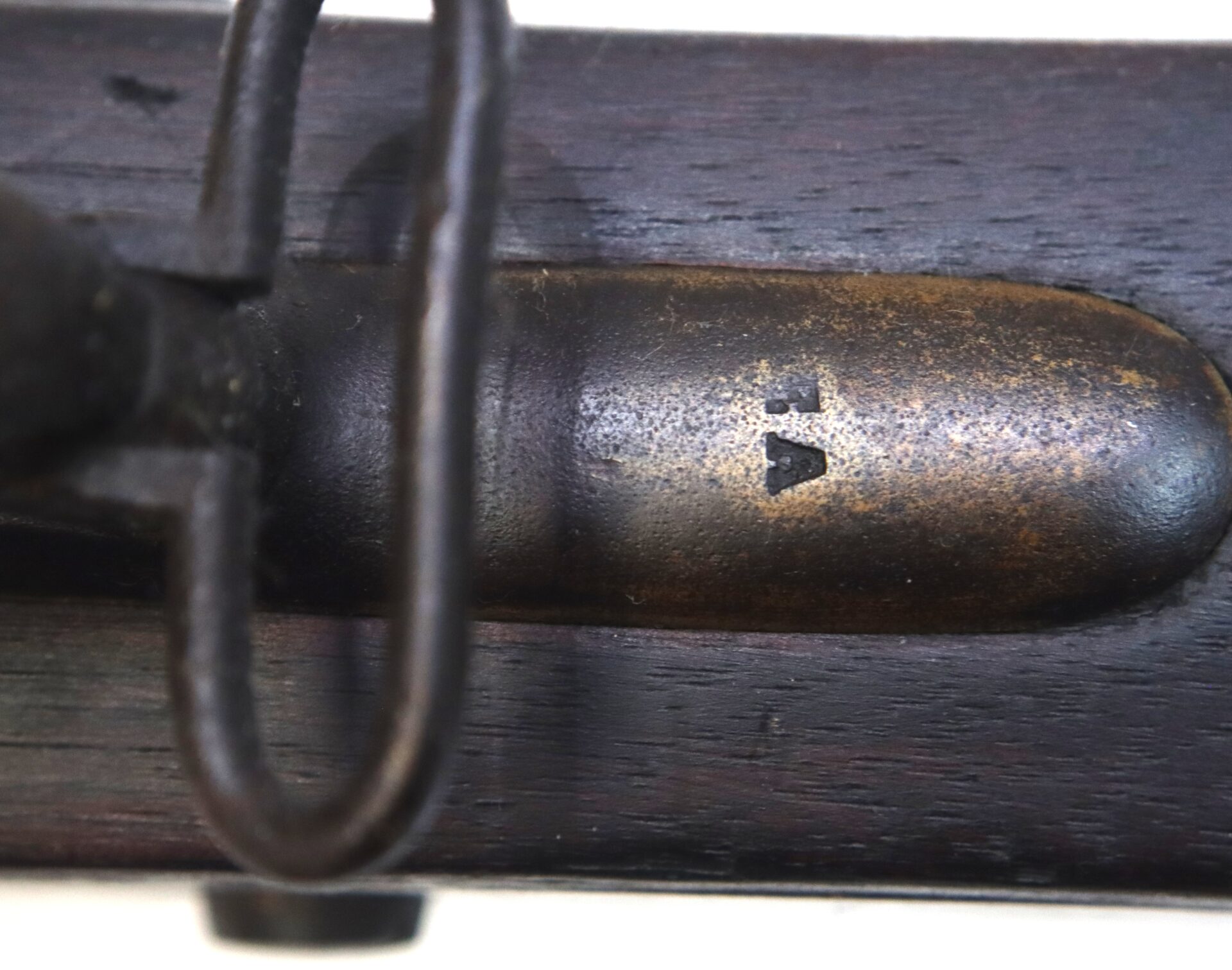 Close-up of a metal and wood surface with a stamped mark resembling a broad arrow pointing upward; partial view of a metal loop is also visible.