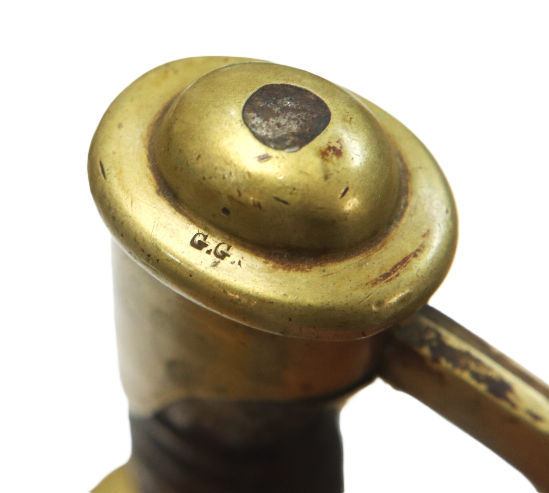 Close-up of a brass object with a rounded cap, showing signs of wear and a stamped marking "G.G." on the surface.