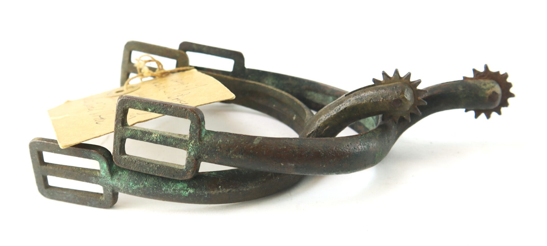 A pair of old metal spurs with rowels, showing signs of tarnish and patina, with a paper tag tied to one.