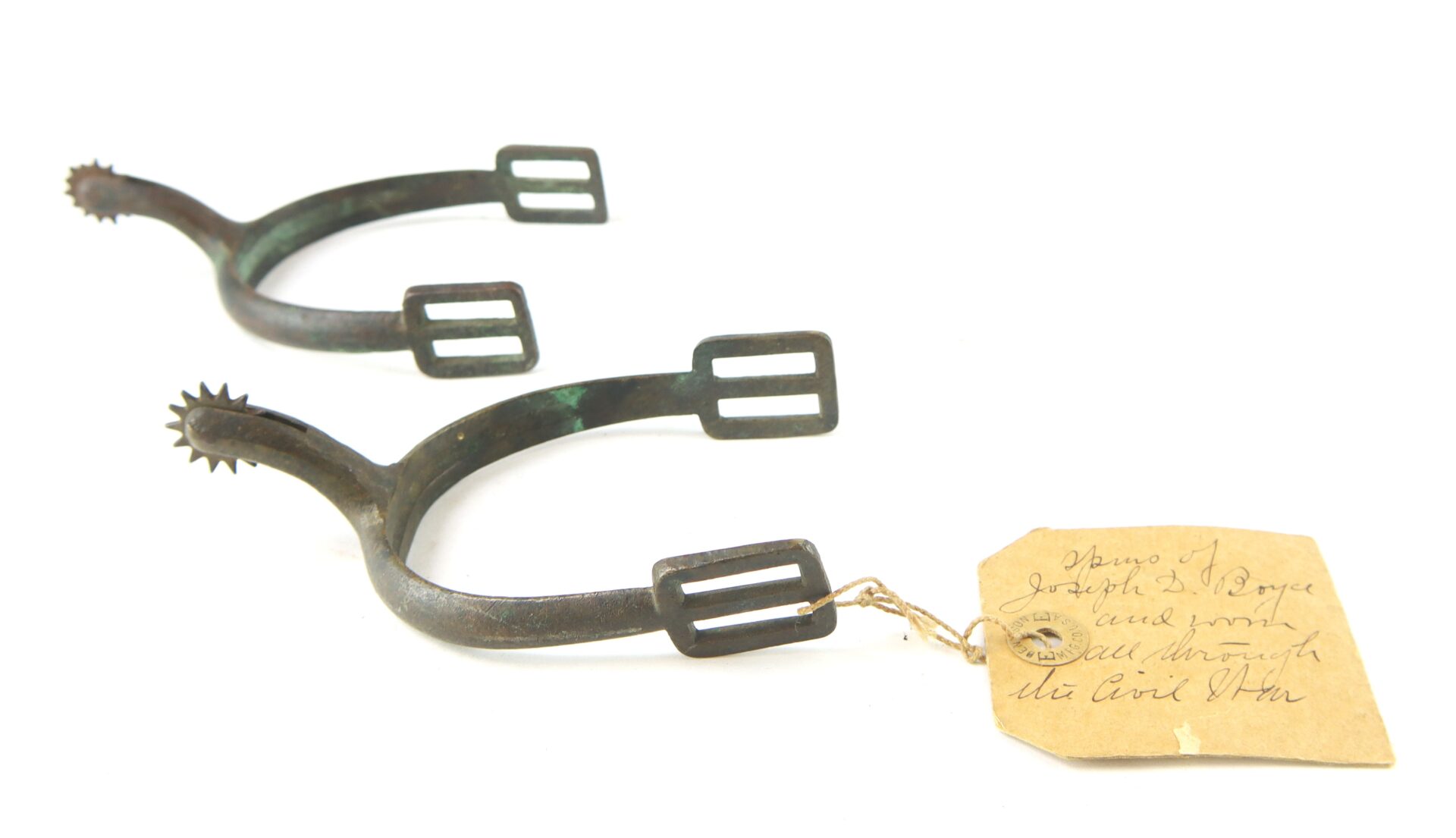 A pair of antique metal spurs with rowels, featuring rectangular buckles and an attached handwritten tag.