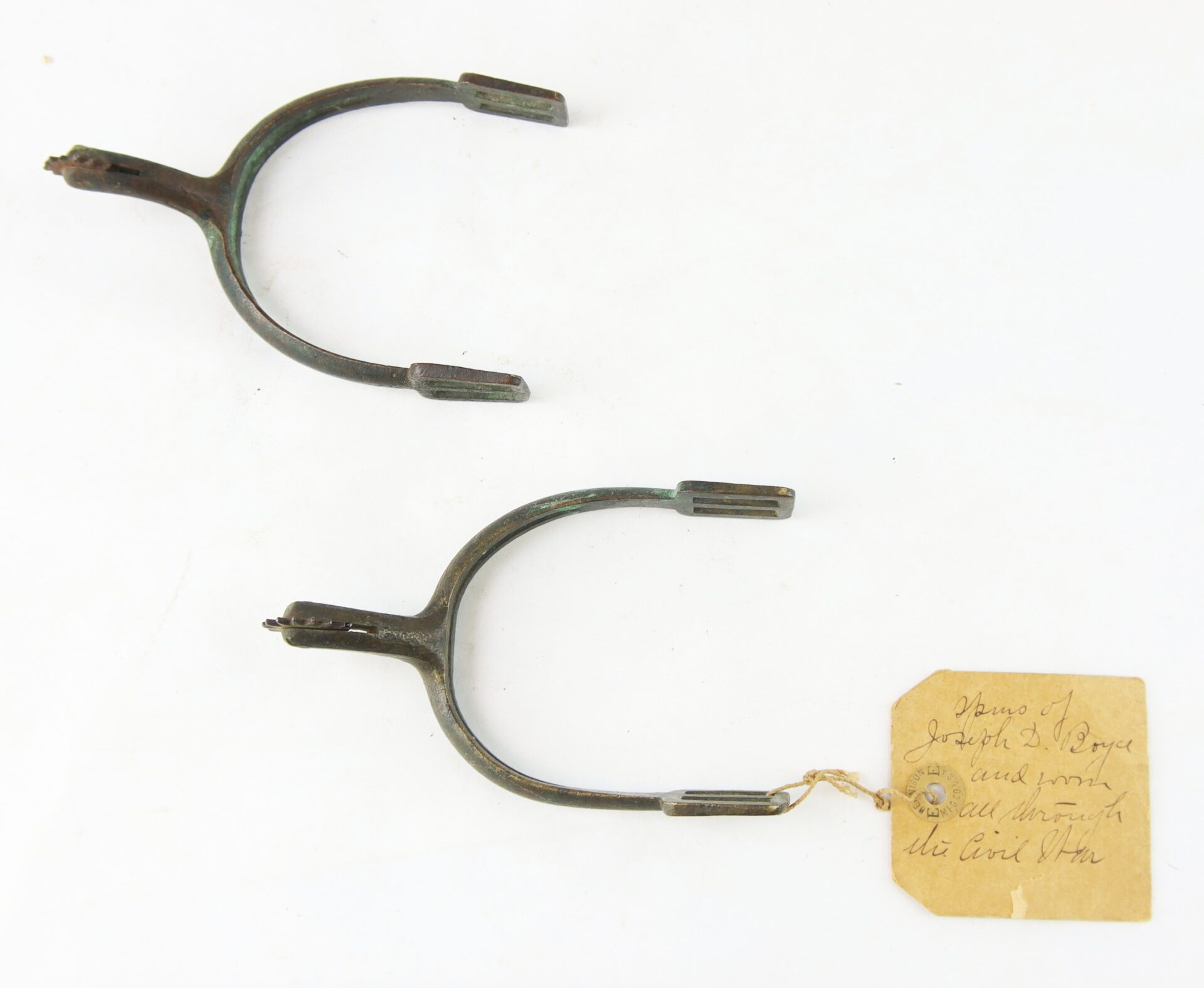 Two antique metal spurs with small rowels, one attached to a handwritten paper tag with string, displayed on a white background.