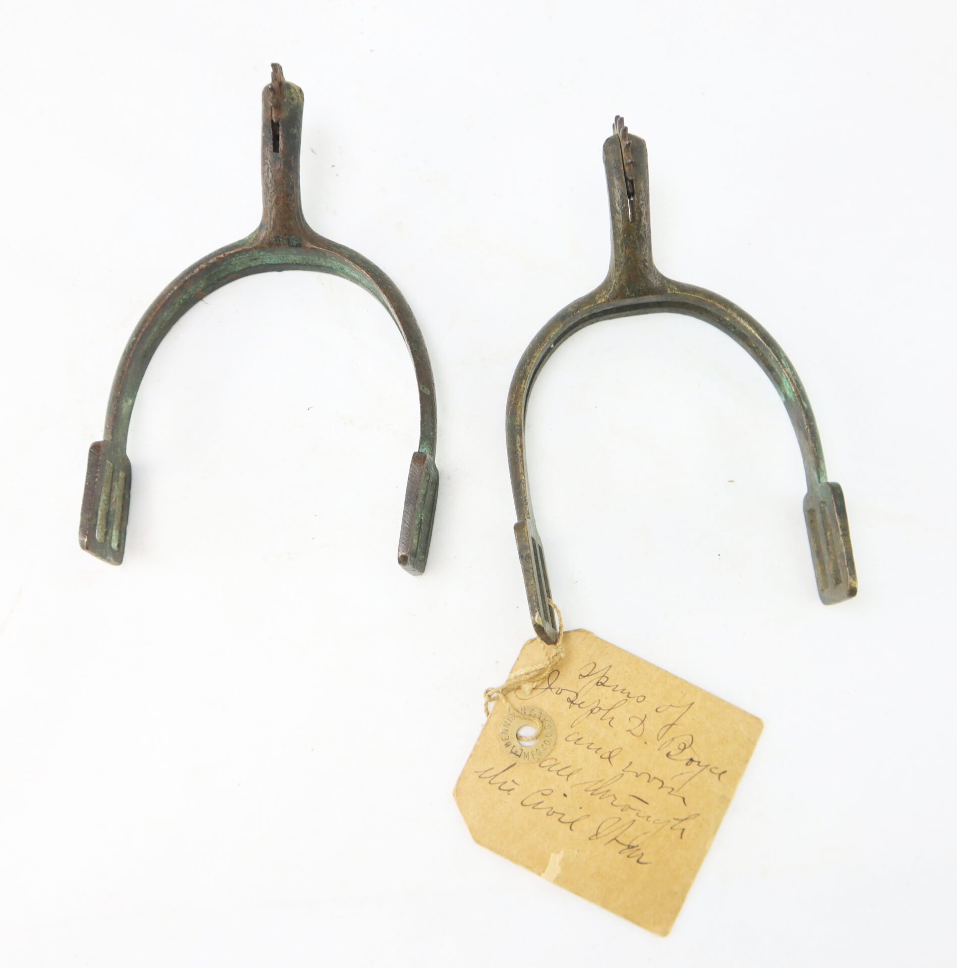 Two old metal spurs with worn greenish patina, one with a handwritten tag attached, placed side by side on a white background.