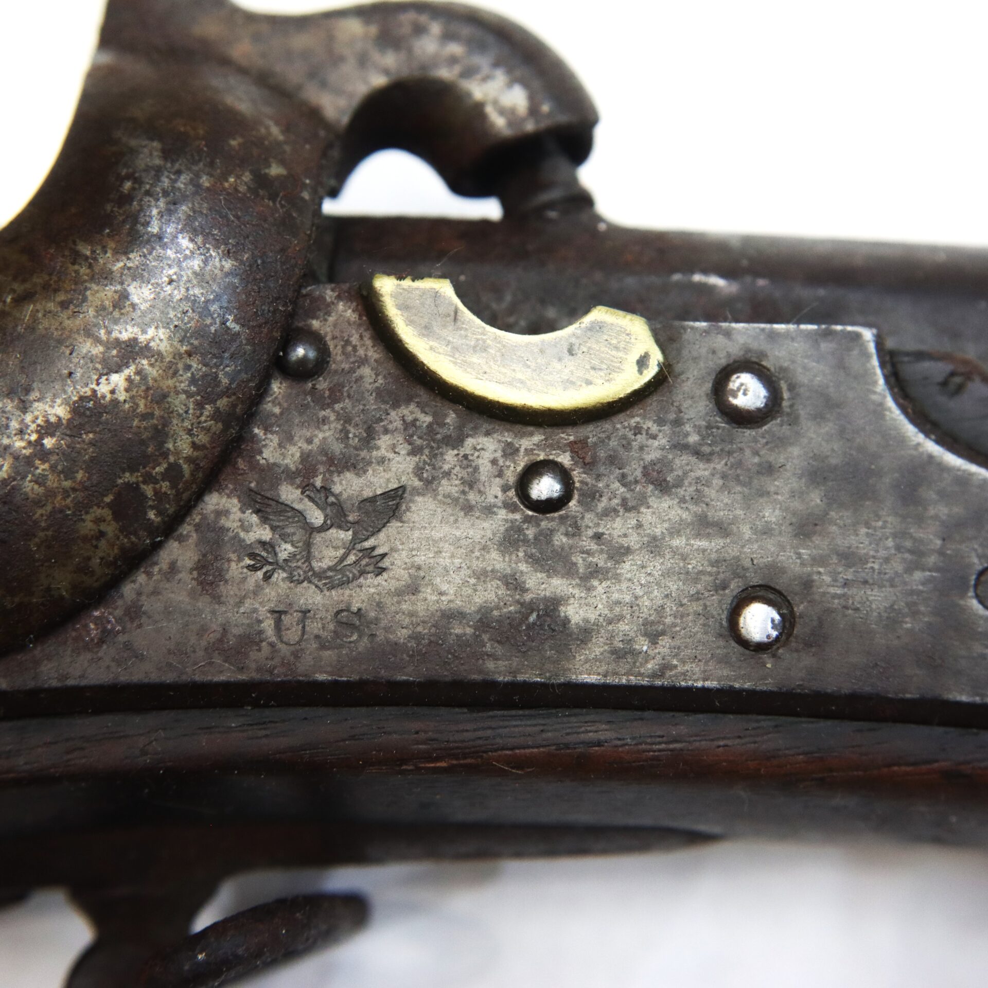 Close-up of an antique firearm's metal receiver, showing a faint U.S. eagle engraving, the letters "U.S.," and several rivets, with some parts exhibiting rust and wear.
