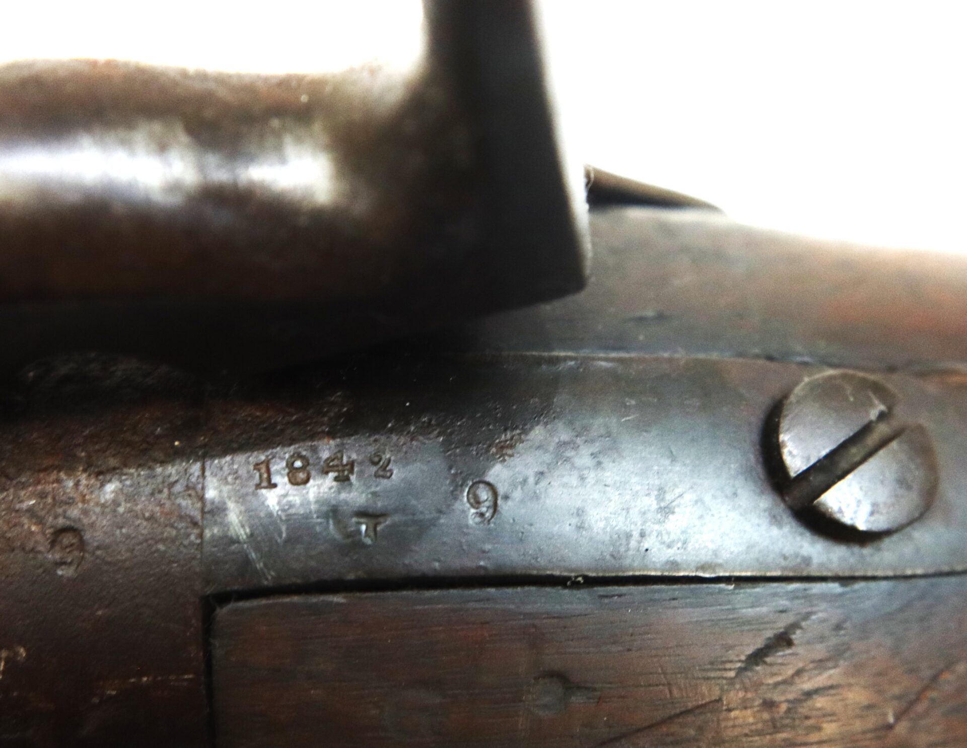 Close-up of an old firearm's metal surface, showing engraved numbers "1842" and a screw, with visible wood grain on the gunstock.