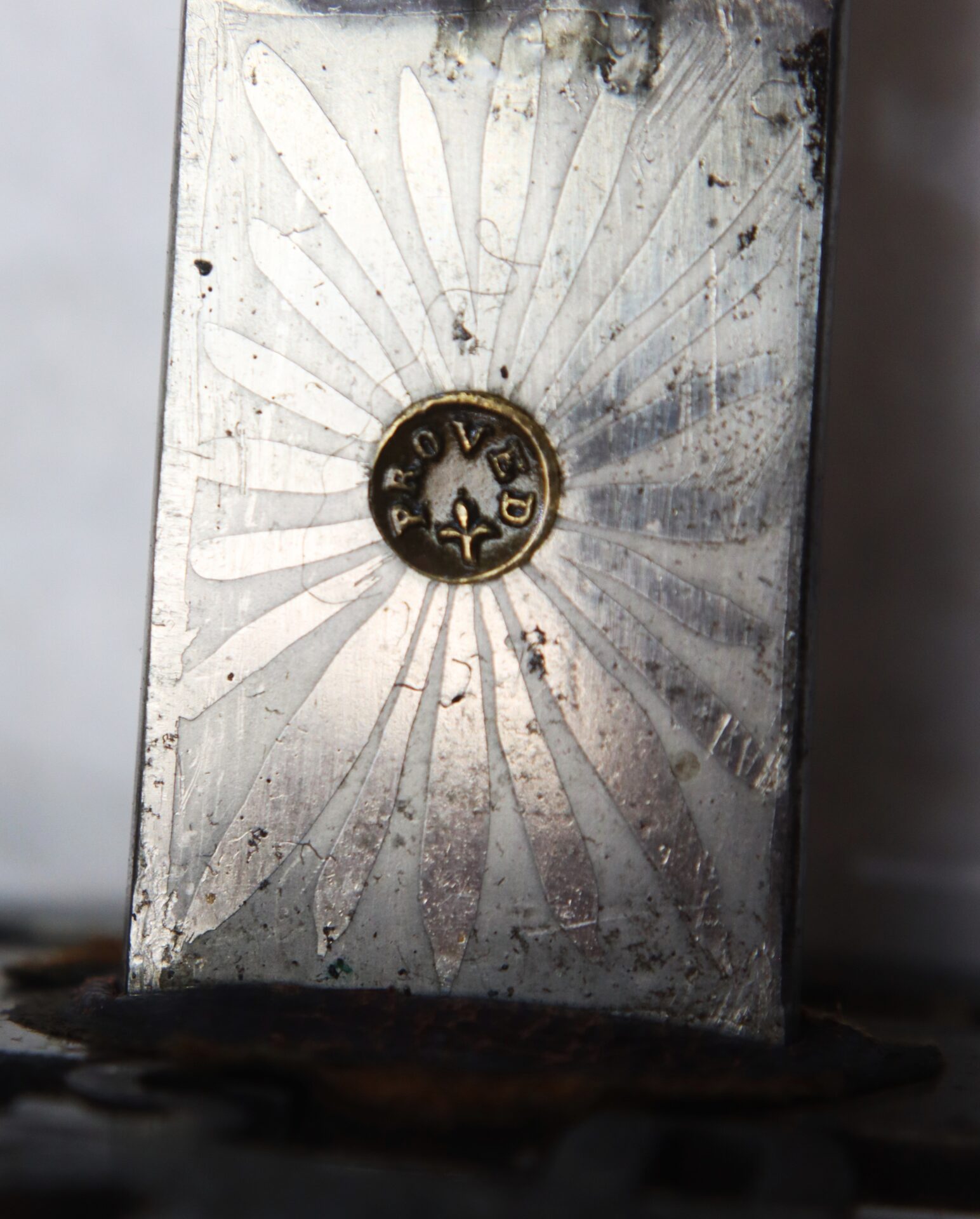 Close-up of a metallic surface engraved with radiating lines and a circular emblem in the center, stamped with the word "APPROVED" and a small fleur-de-lis symbol.
