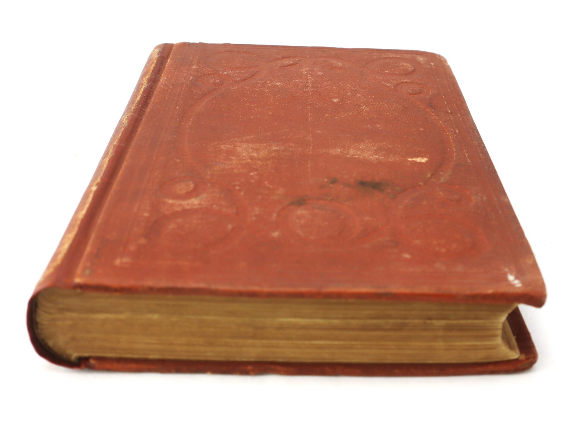 A closed, worn, red hardcover book with embossed decorative patterns, viewed from the side, showing yellowed pages.