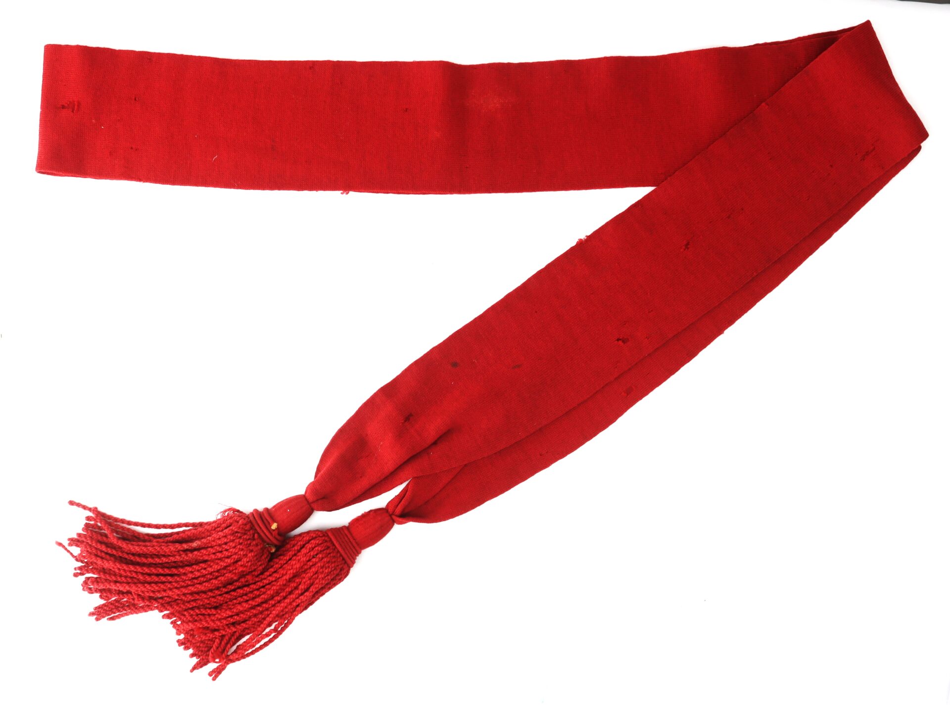 A long, red fabric sash with two tassels at one end, laid out on a white background.
