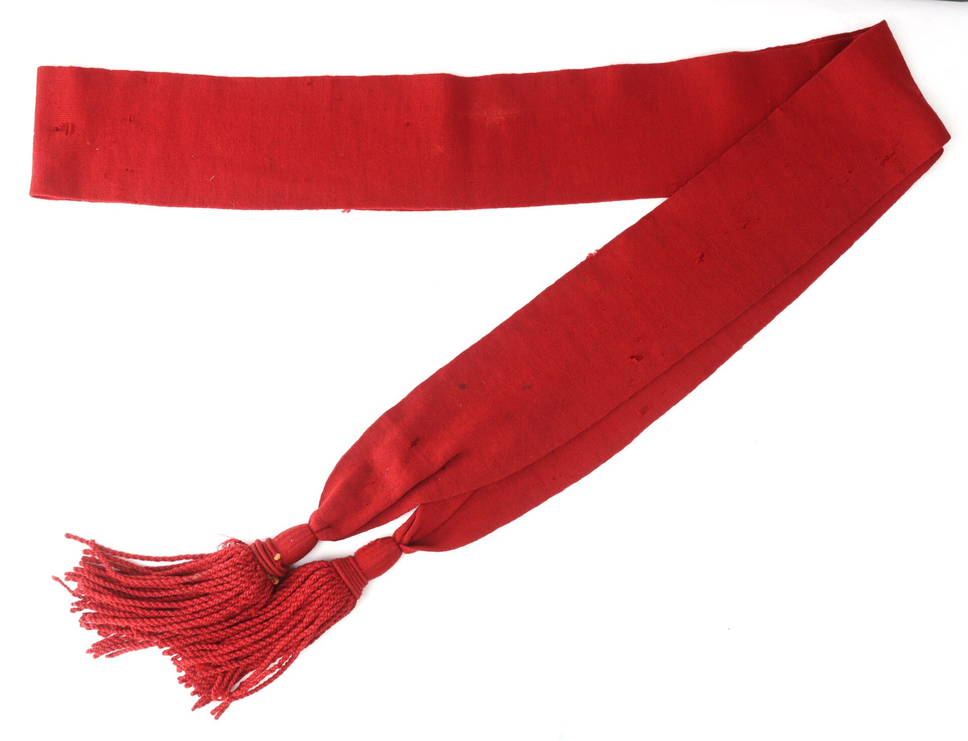 A long, red sash with two tassels at one end, laid flat against a white background.