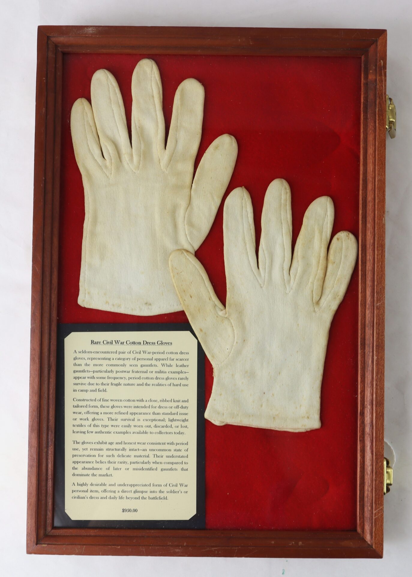 A pair of white cotton gloves displayed in a wooden case with a red background and an informational plaque below them.