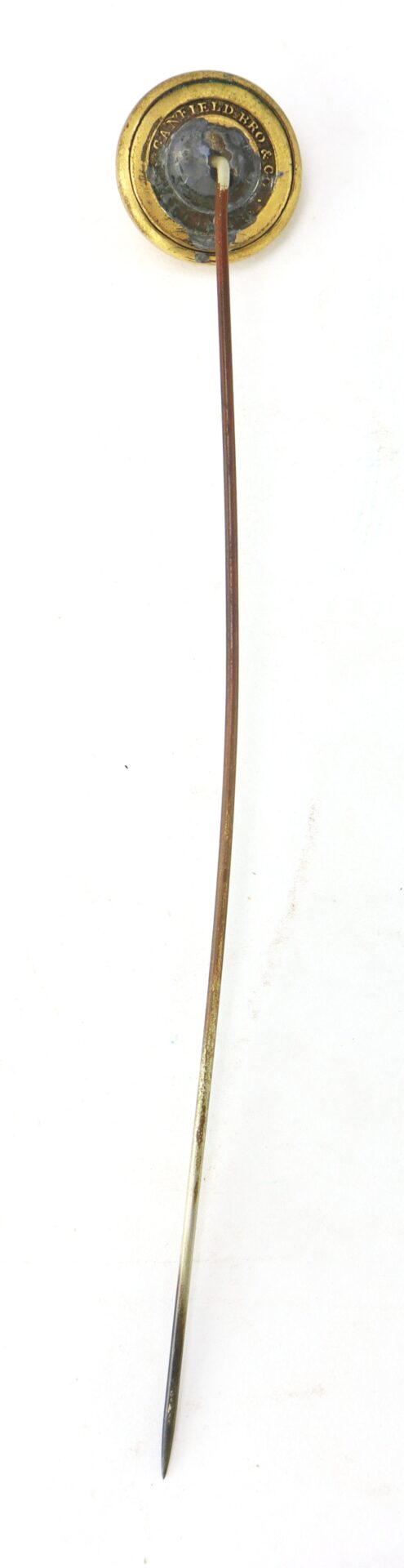A long, thin metal stick pin with a round, gold and black decorative top, viewed against a white background.