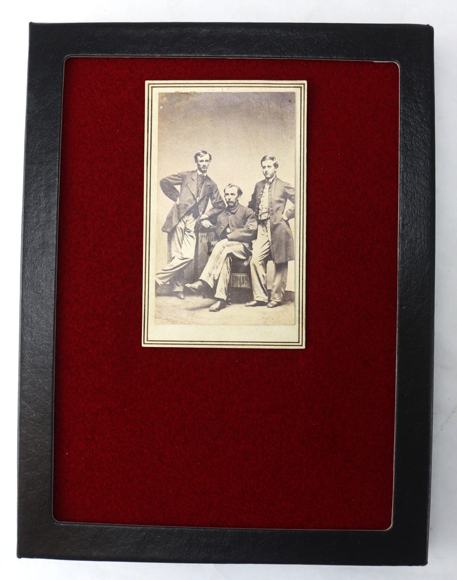 A sepia photograph of three men in 19th-century attire, mounted on a red felt background in a black frame.
