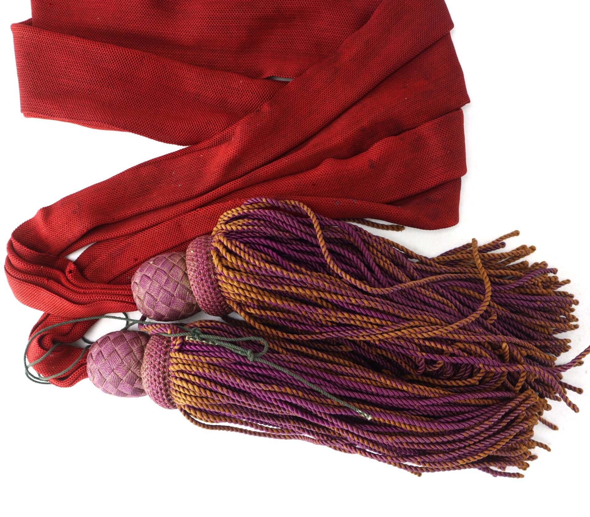 A red fabric sash paired with two purple and gold decorative tassels on a white background.