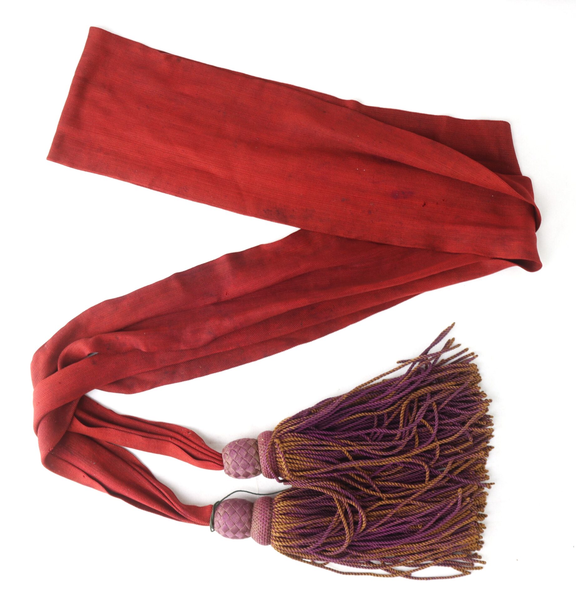 A long, red fabric sash with two purple tasseled ends, featuring decorative knotting near the tassels, placed on a white background.