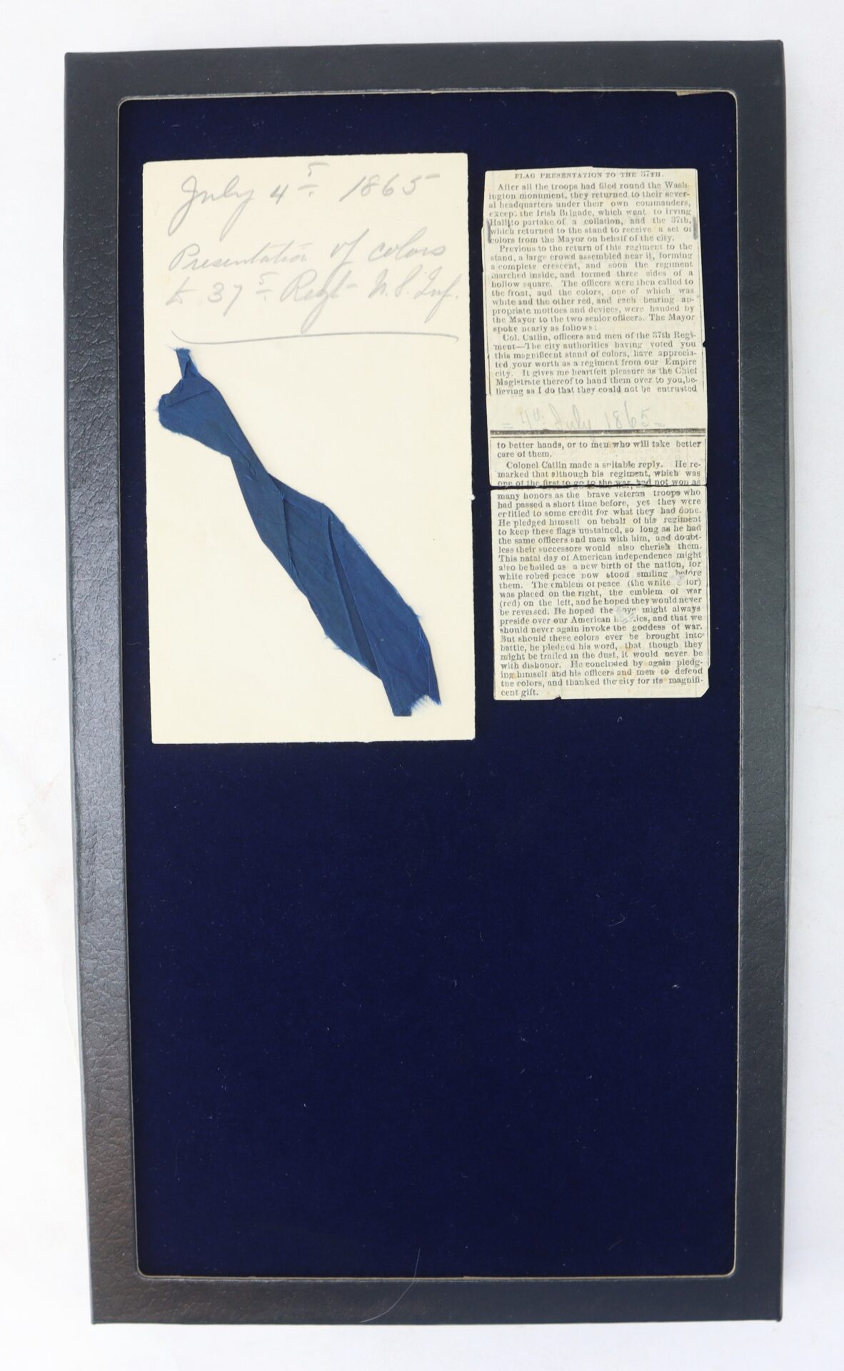 A framed display featuring a handwritten note dated July 4, 1865, a blue fabric ribbon, and two newspaper clippings, all mounted on a dark blue background.