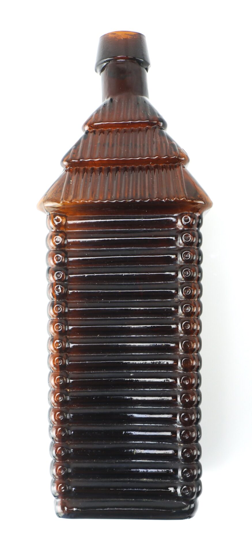 A brown glass bottle shaped like a log cabin, with ribbed sides and a ridged roof design.