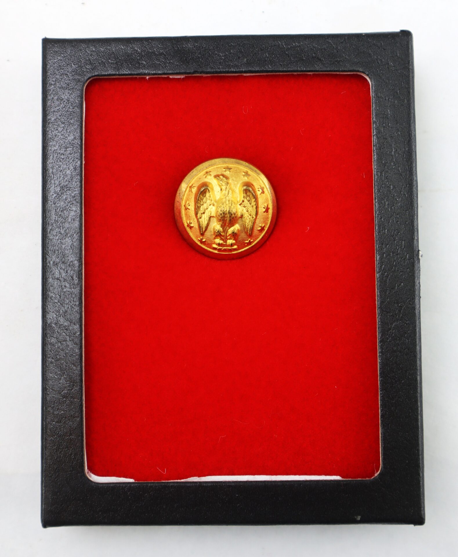 A gold button with an eagle emblem displayed in a black frame with a red velvet background.