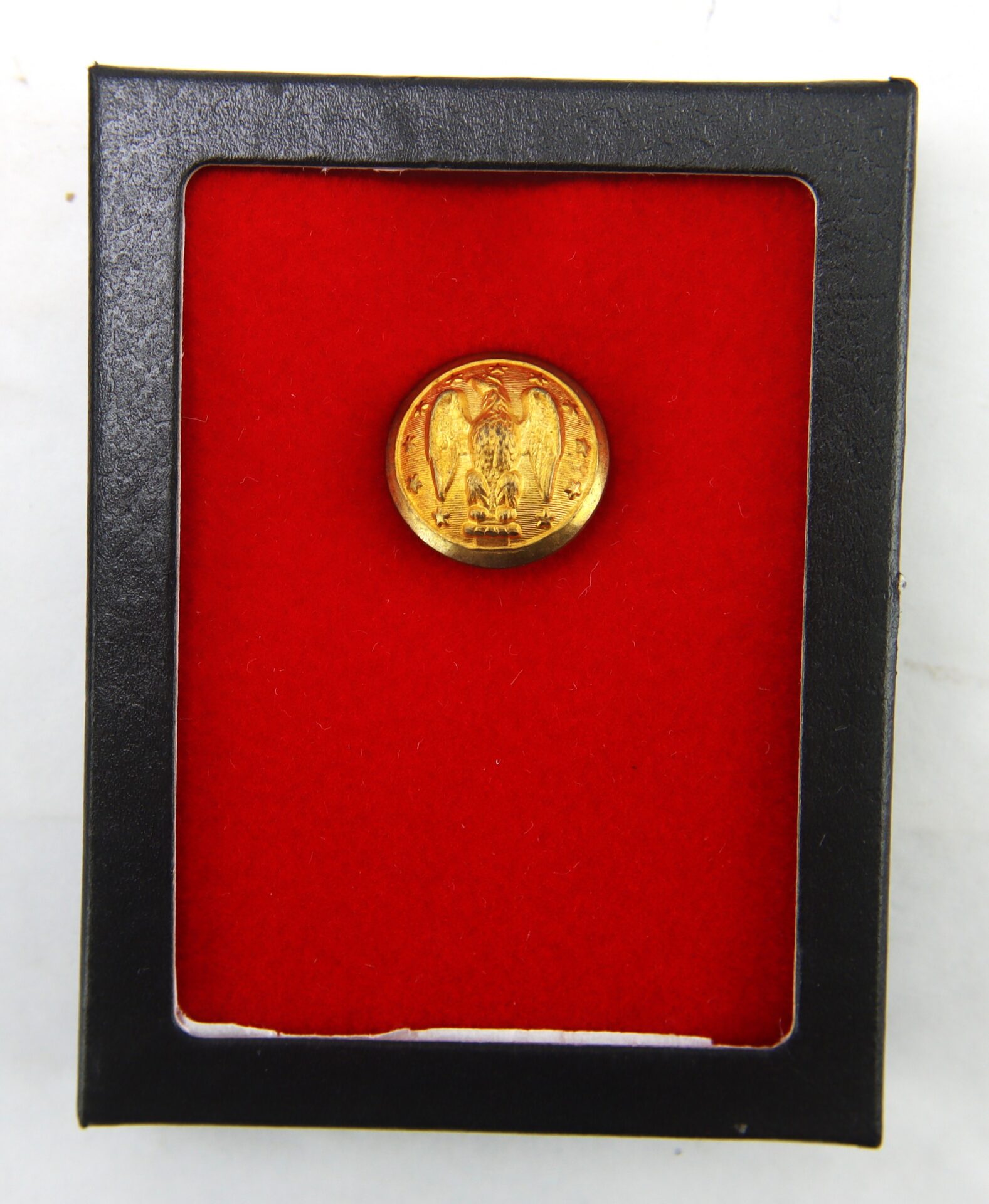 A gold button with an eagle design is displayed in a black case lined with red velvet.