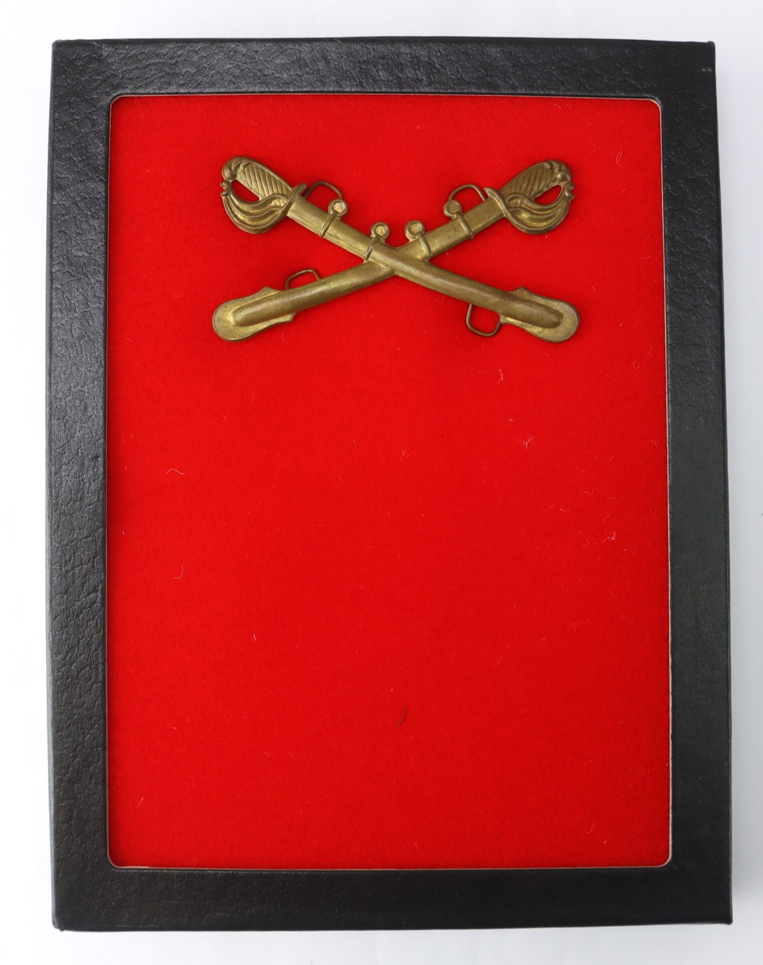 Crossed brass cavalry sabers insignia mounted on a red felt background inside a black rectangular frame.