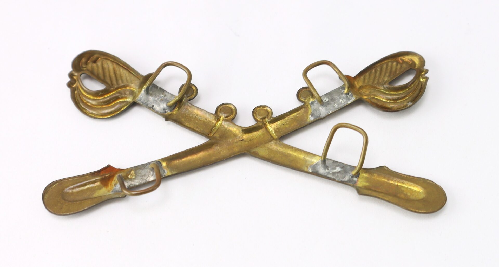 A brass crossed sabers insignia with three attachment loops on each sword, placed on a white background.