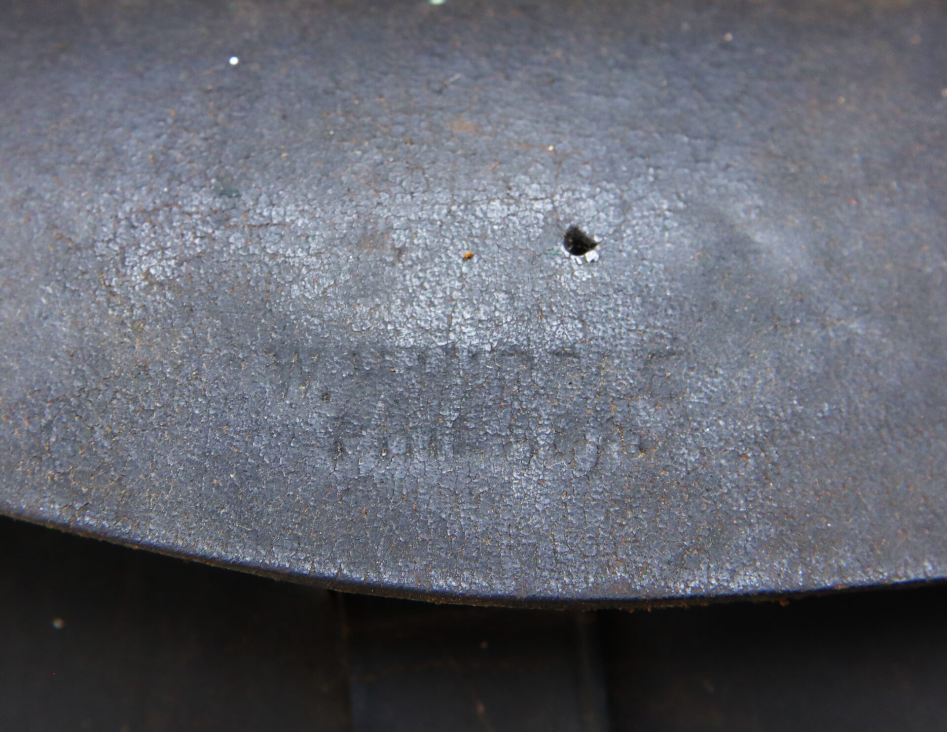 A close-up of a rusted, metal surface with weathered, barely visible stamped markings and a small hole near the center.