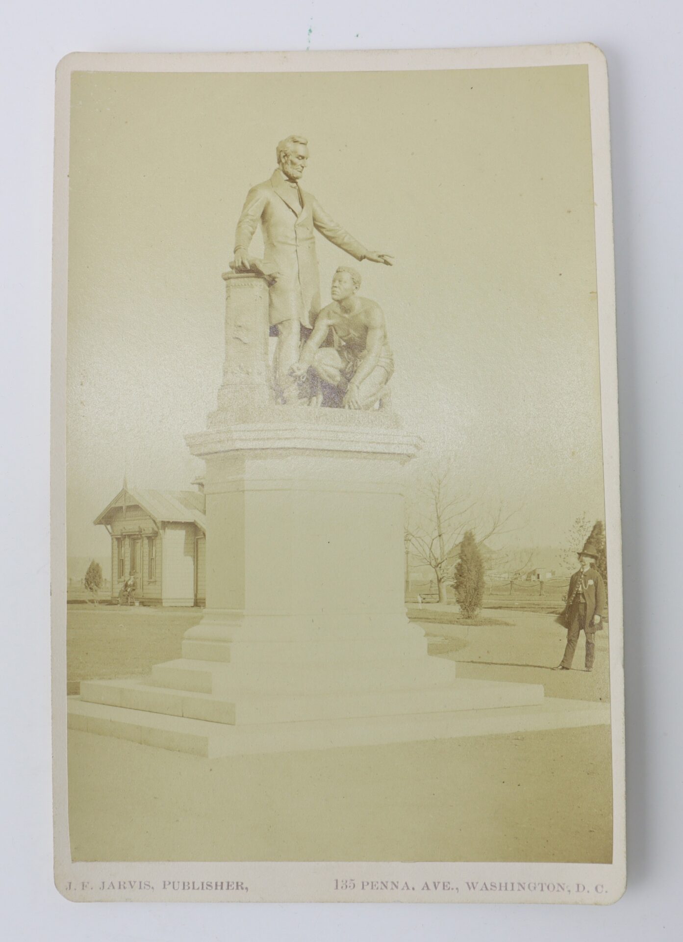 Sepia-toned photo of a statue featuring Abraham Lincoln standing beside a kneeling, shirtless man in shackles on a large pedestal, with buildings and trees in the background.