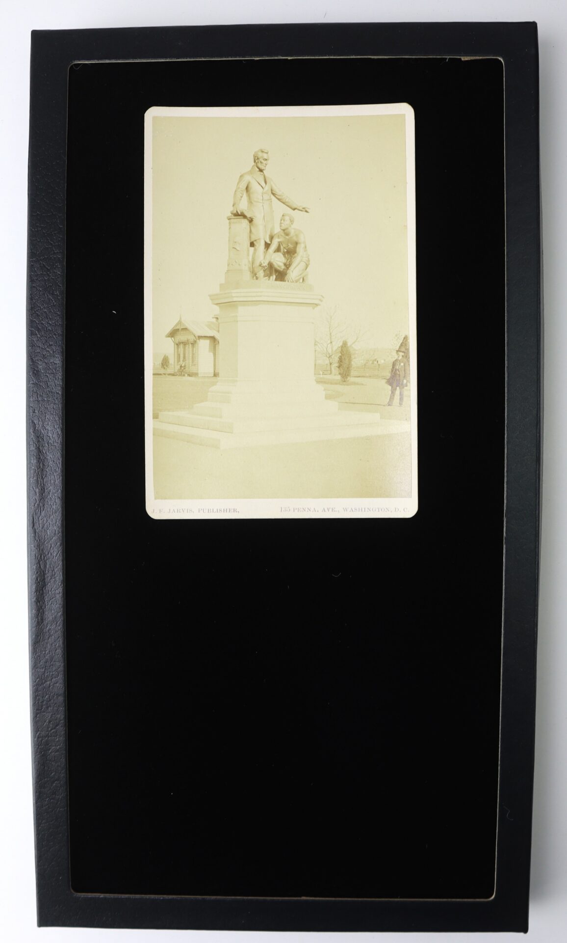 Sepia photograph of a statue showing one standing and one kneeling figure on a pedestal, mounted on a black mat with a domed building visible in the background.