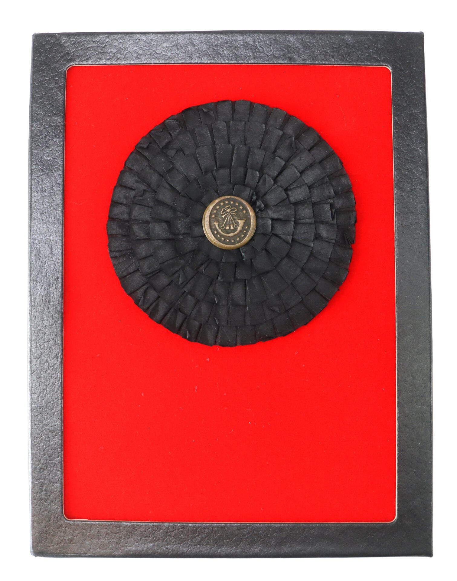 A black rosette with a circular emblem at the center is mounted on a red background within a rectangular black frame.