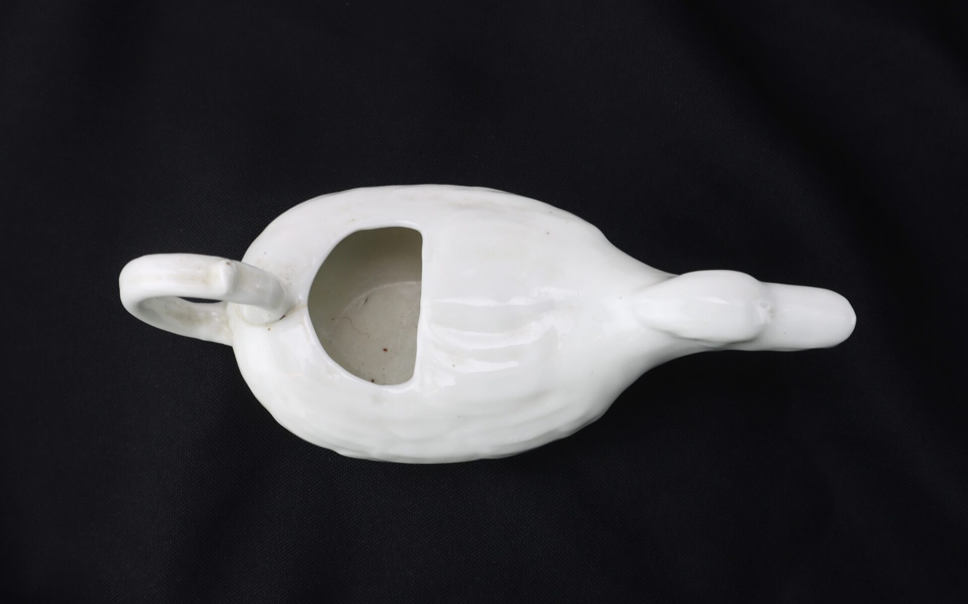 A white ceramic gravy boat shaped like a goose, viewed from above, showing an open top and handle, on a black background.
