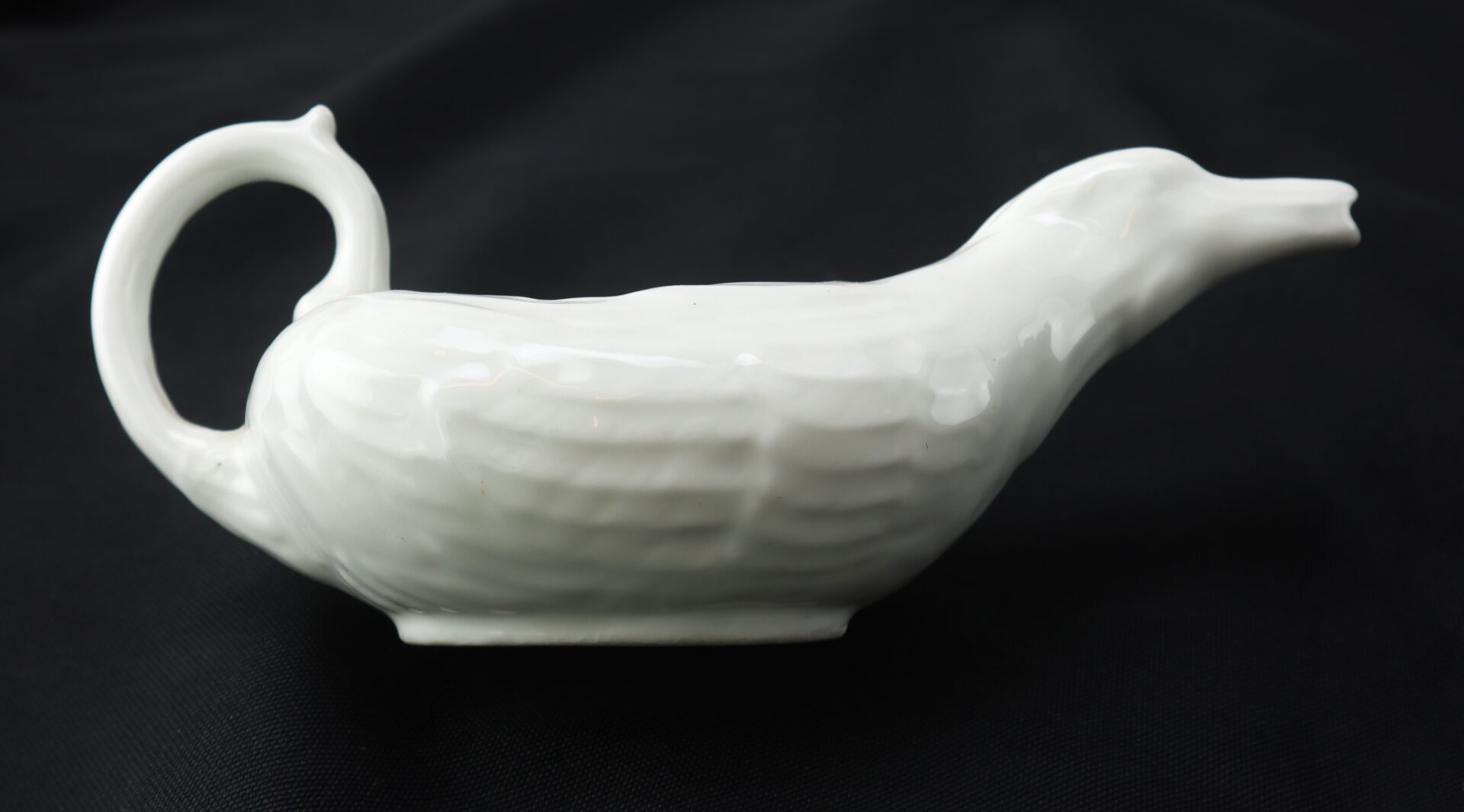 White ceramic gravy boat shaped like a duck with a textured body, curved handle at the tail, and spout at the beak, set against a black background.