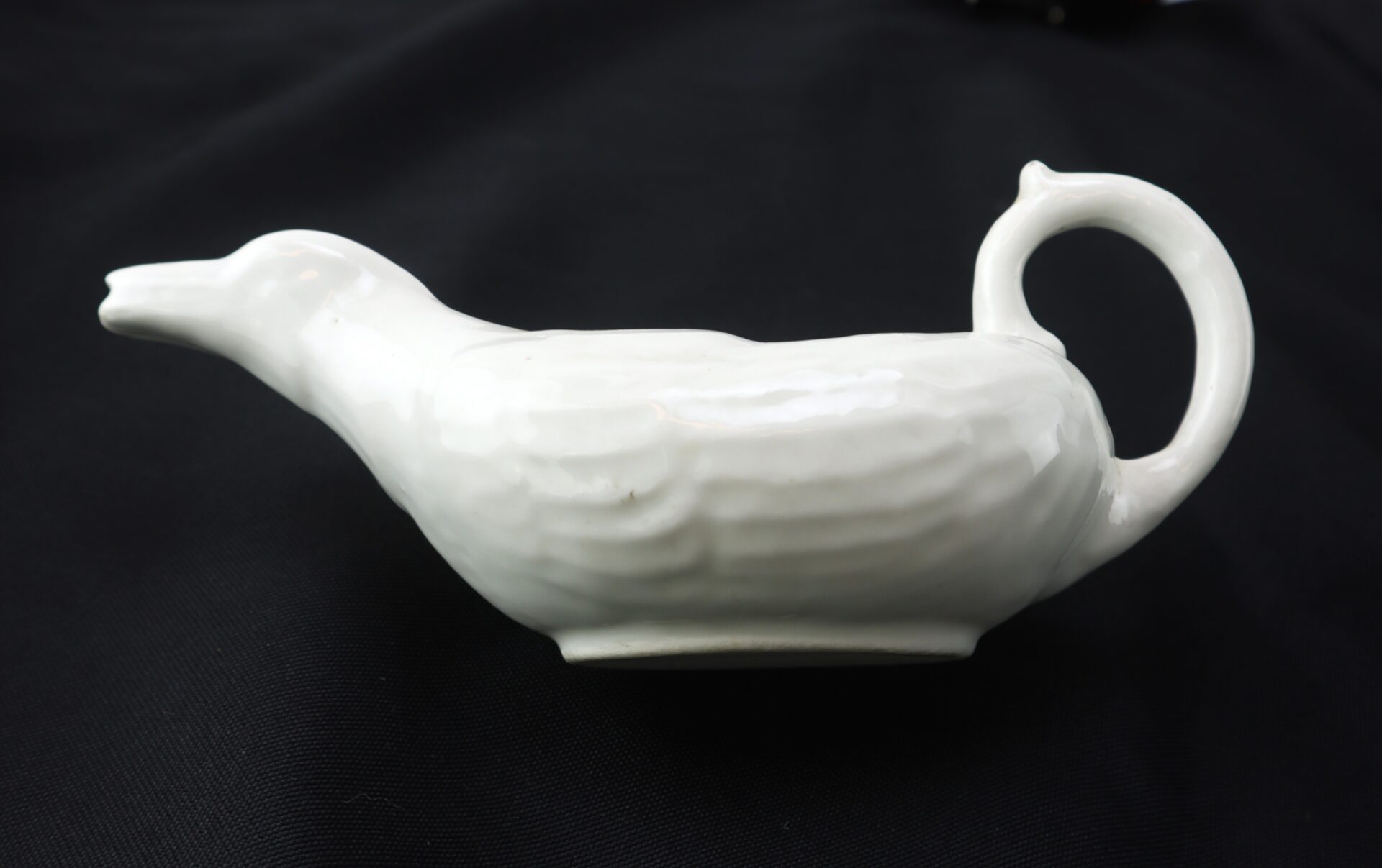 White ceramic gravy boat shaped like a bird, with a handle resembling a curled tail, placed on a black background.