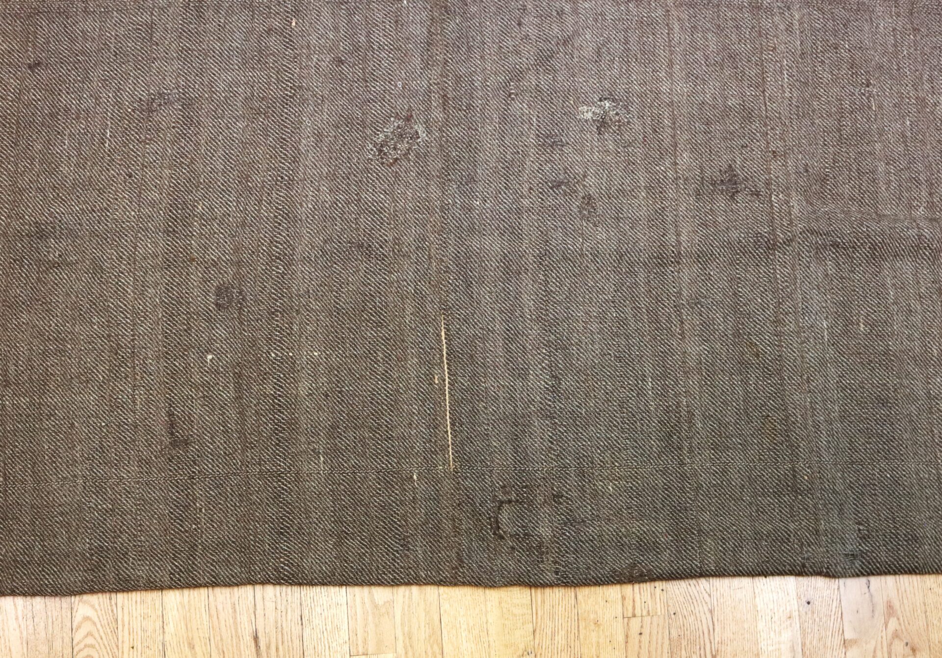 A brown woven rug with visible wear, stains, and patches, lying on a light wooden floor.