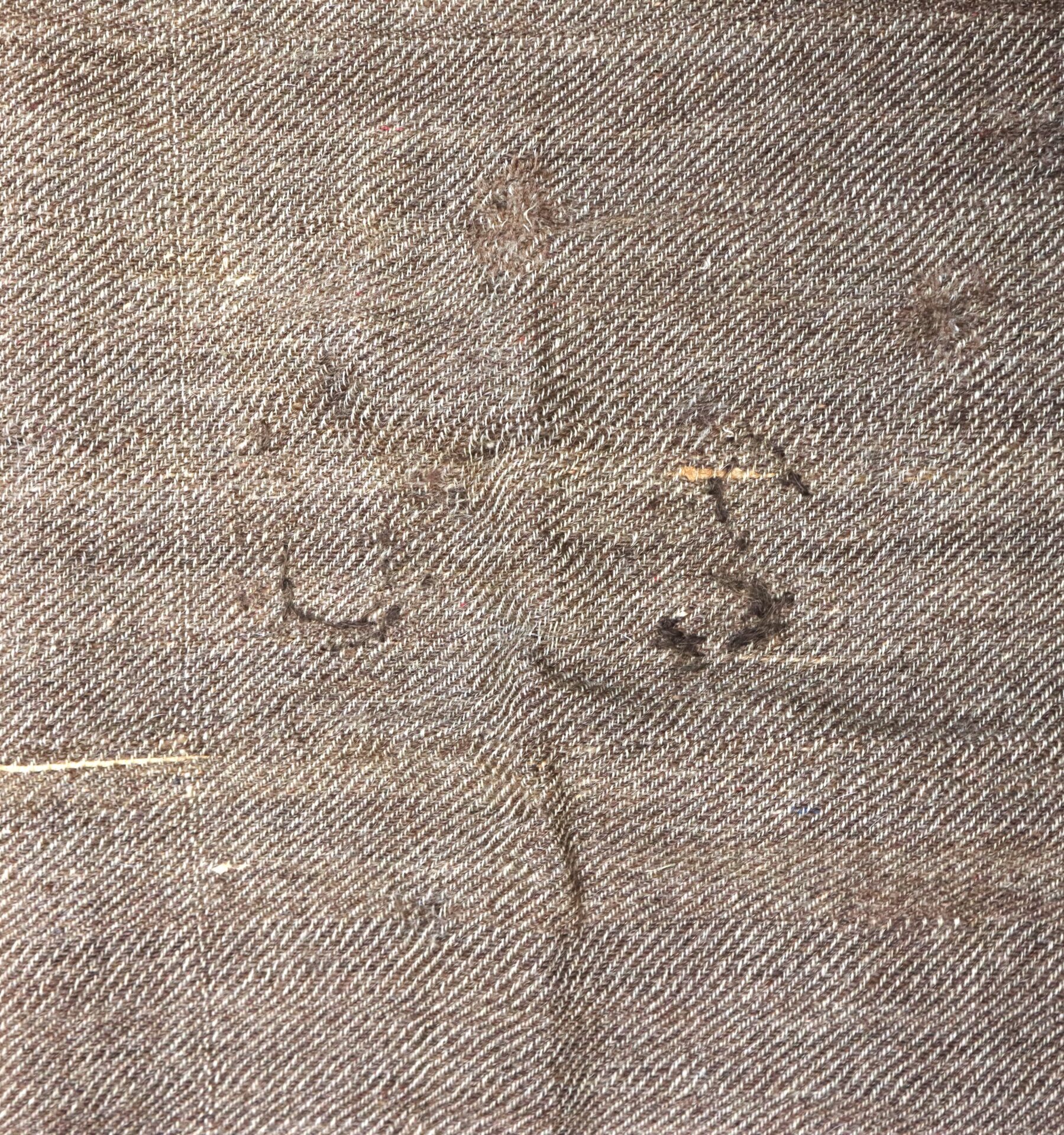 Close-up of a textured, worn fabric in a brownish tone, with visible stitches, seams, and some fraying thread.