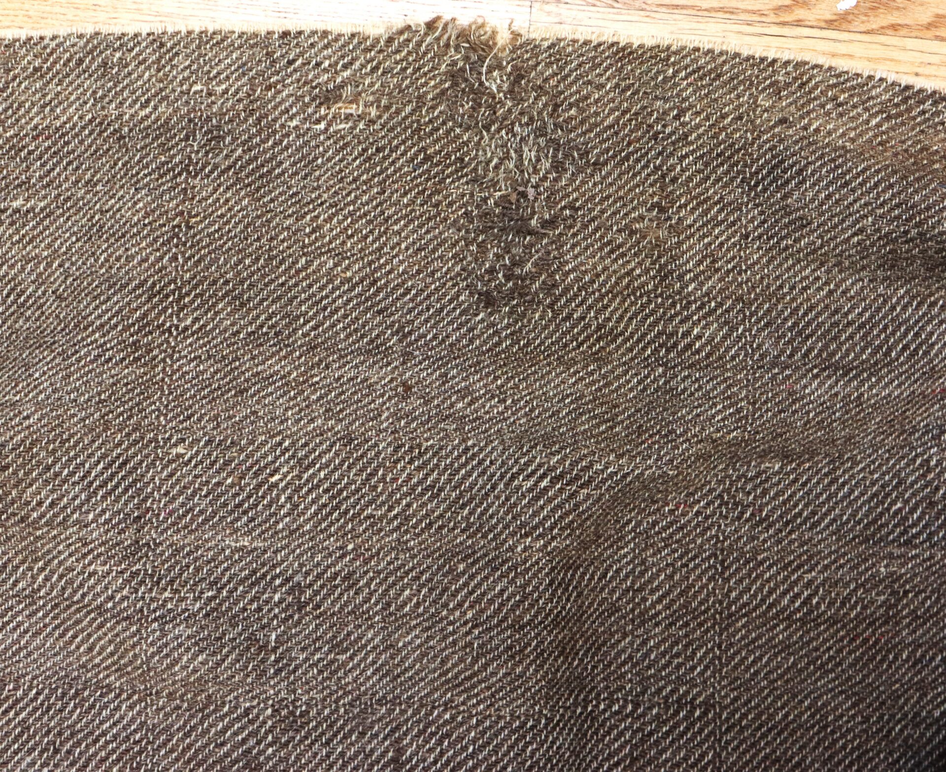 Close-up of a brown tweed fabric with a visible worn patch and frayed threads near the top center, lying on a wooden surface.
