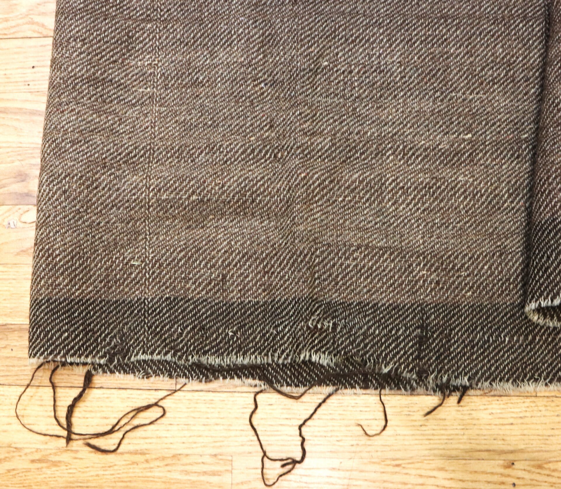 A close-up of a brown woven fabric with frayed edges and loose threads, laid on a wooden floor.