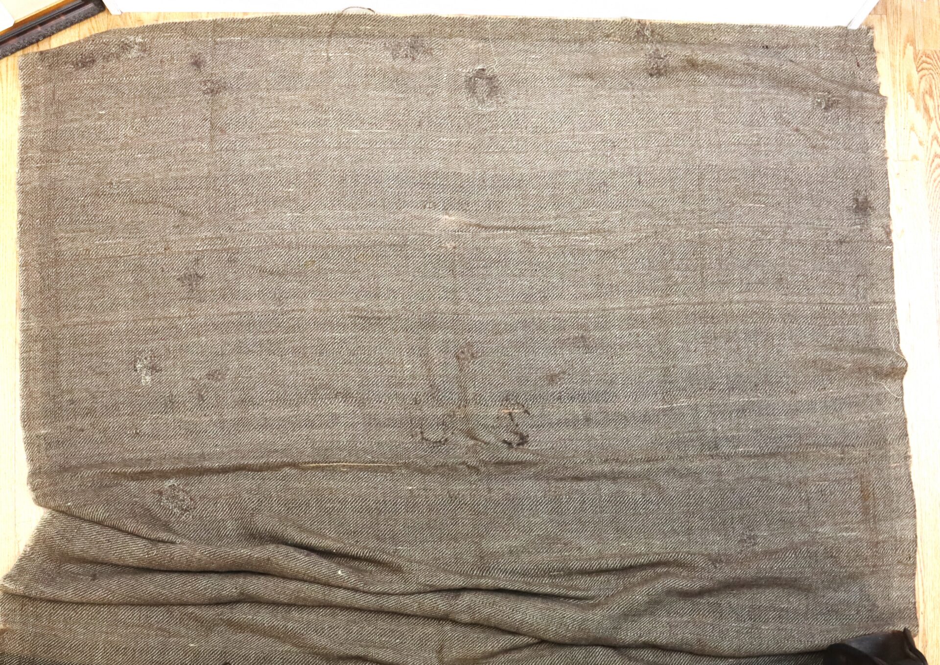 A large, worn piece of brownish fabric with visible patches, wrinkles, and frayed thread, laid out on a wooden surface.