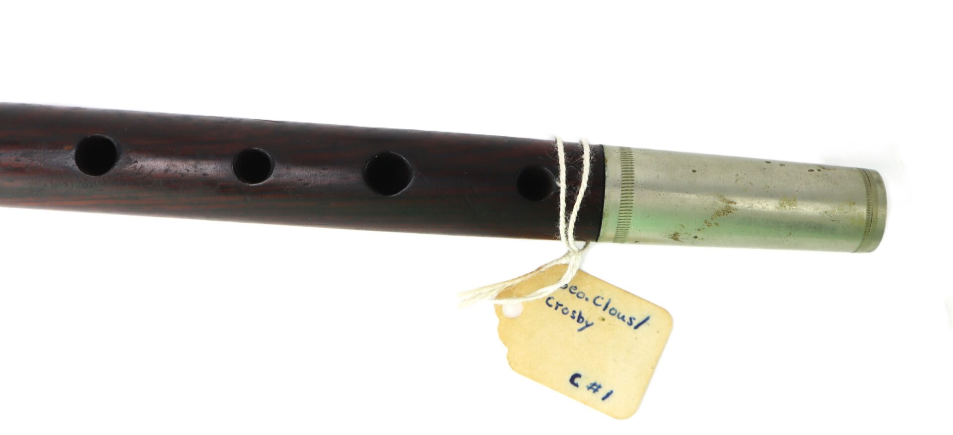 A close-up of a wooden flute or fife with a metallic end, showing finger holes and a string-attached tag labeled "Geo. Cloasf Crosby C41.