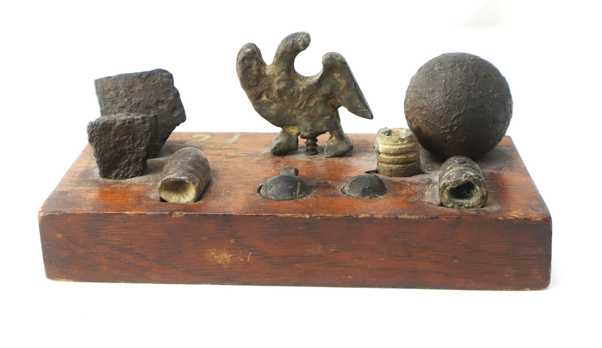 A wooden base displaying various metal relics, including a round ball, an eagle figure, chunks of metal, and cylindrical objects.