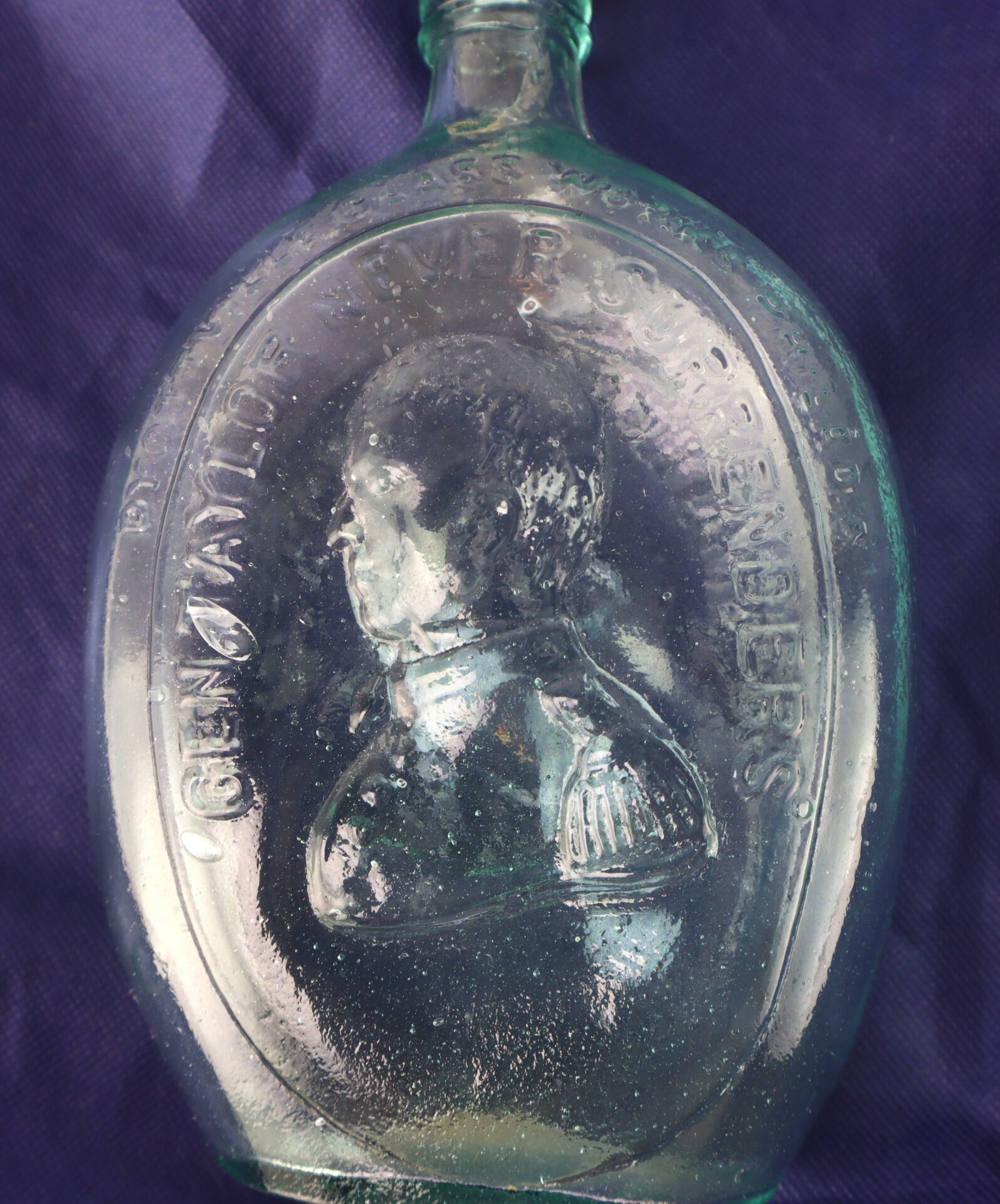 A clear glass bottle embossed with a profile portrait of a man in military uniform and the words "GEN. TAYLOR NEVER SURRENDERS" against a dark blue fabric background.