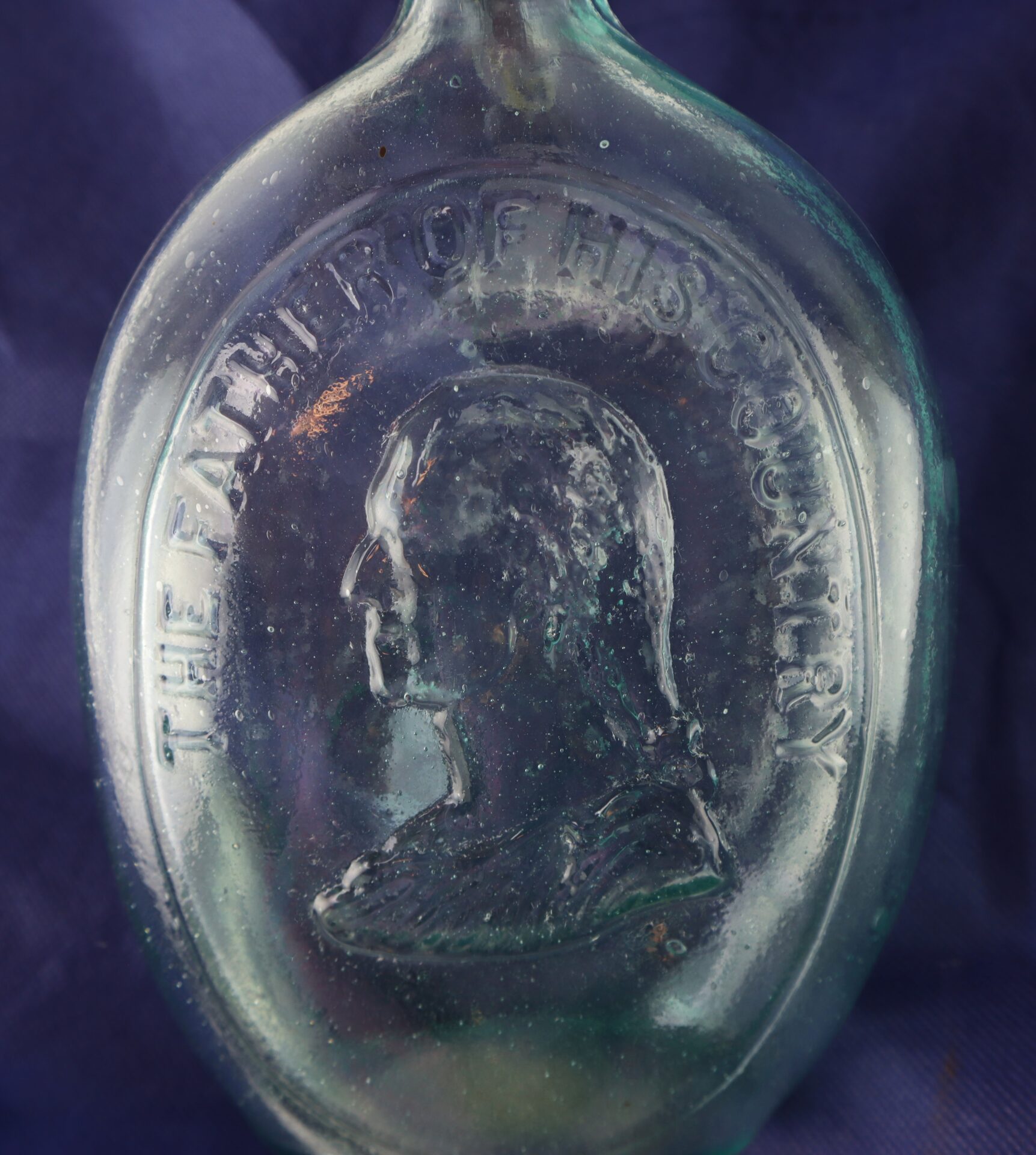 A close-up of an old glass bottle featuring an embossed profile of a man with the words "THE FATHER OF HIS COUNTRY" around the portrait.