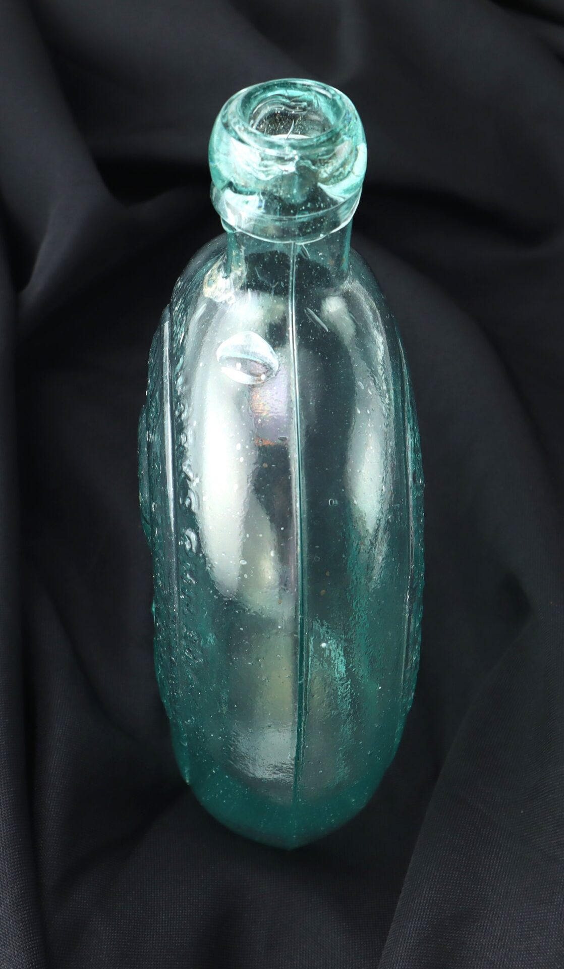 A vintage, light blue-green glass bottle with vertical ridges is placed upright on a black fabric background.