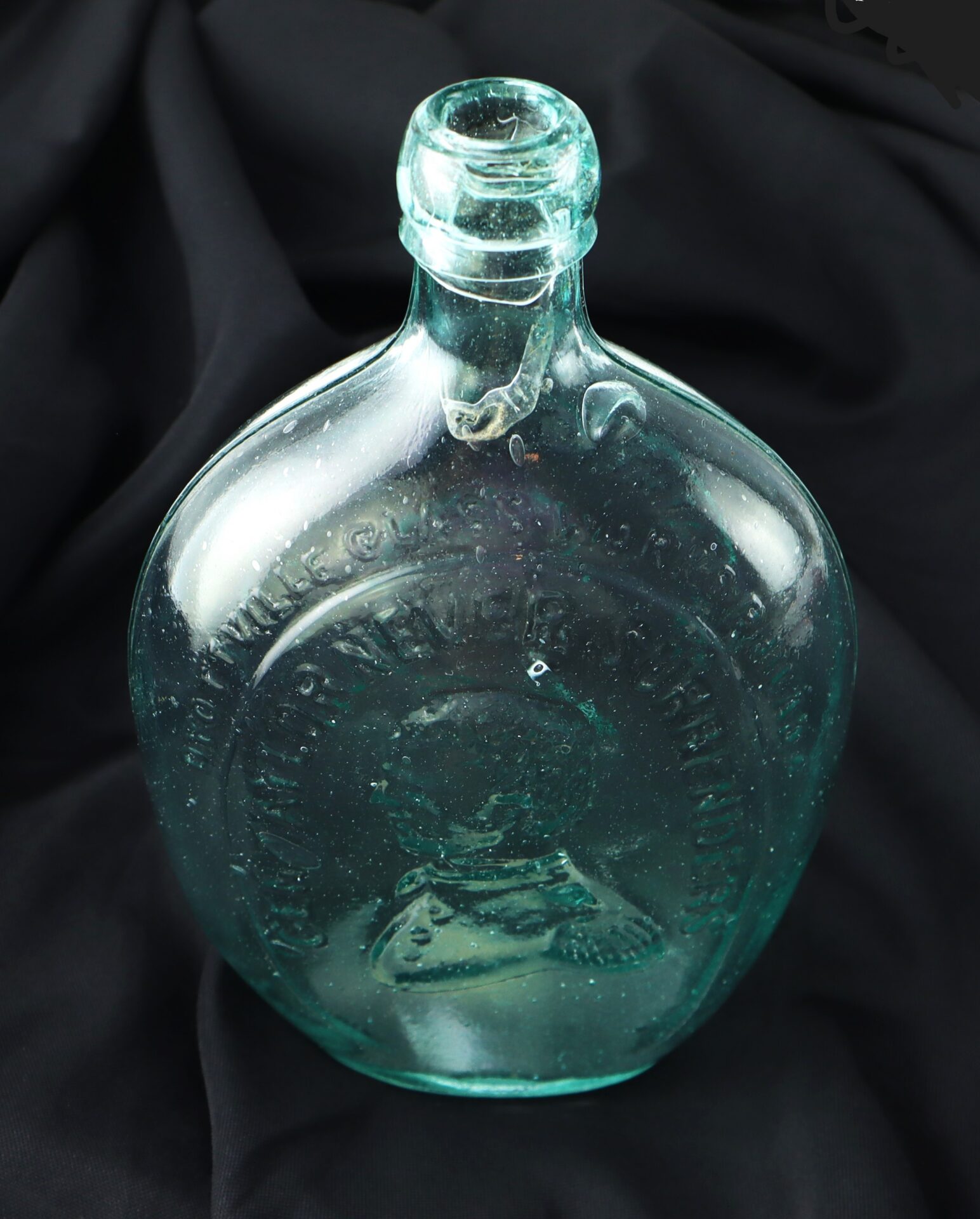 A vintage, clear glass bottle with embossed text and an embossed portrait on one side, set against a black fabric background.