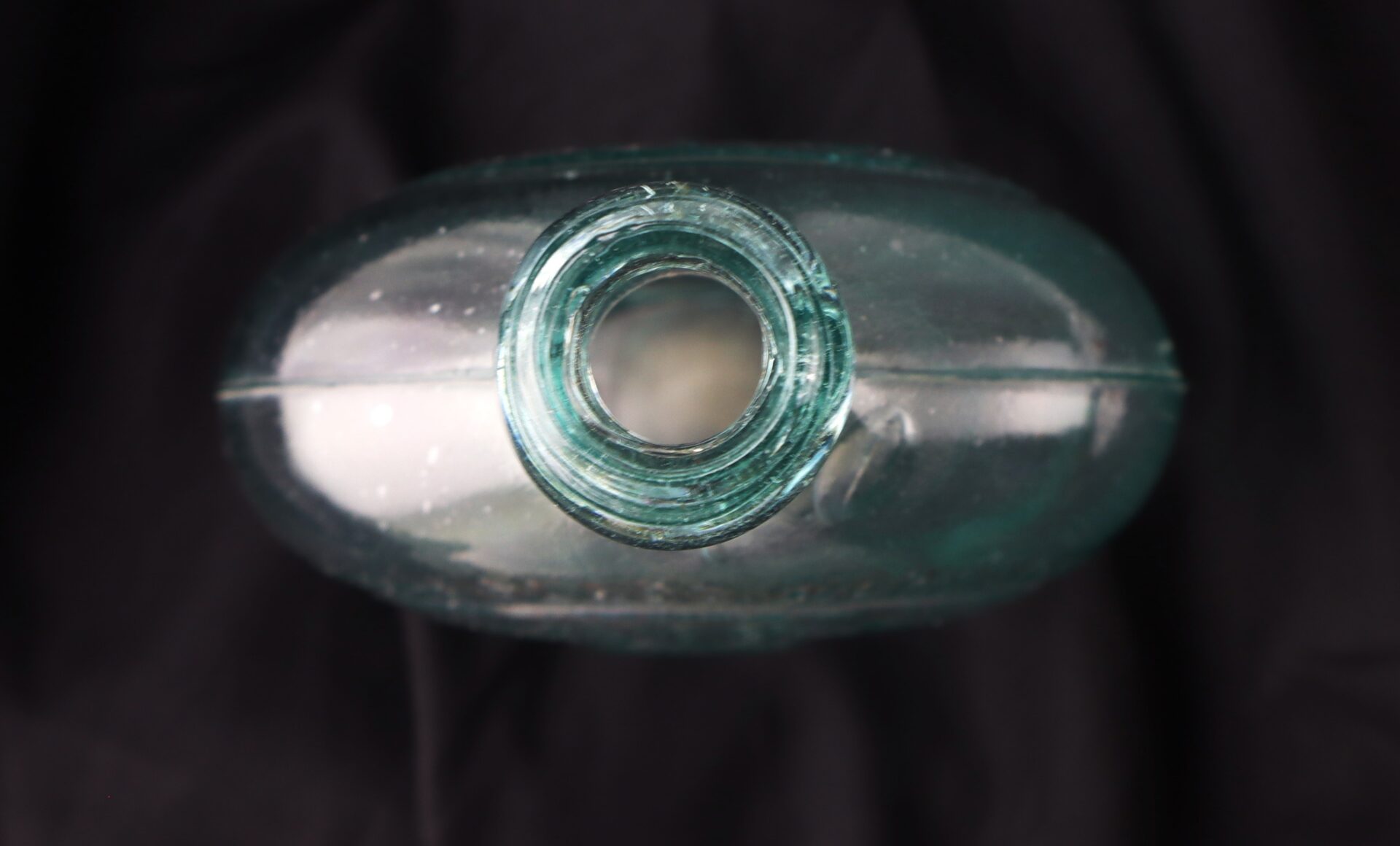 Top down view of an empty, clear glass bottle with a wide opening, placed on a black fabric background.