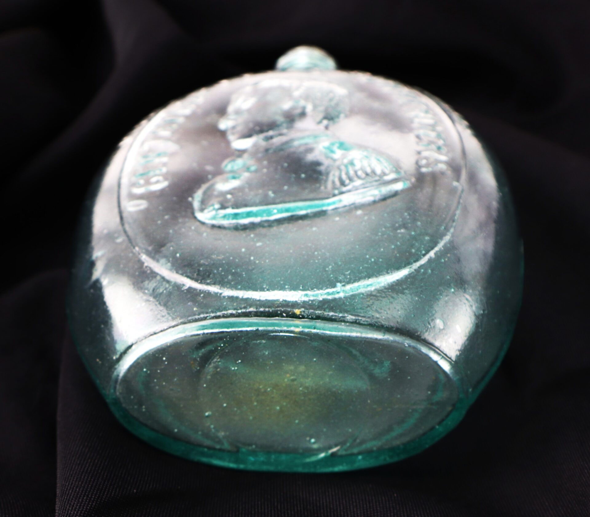 A vintage blue-green glass bottle with an embossed portrait of a man in profile and raised text, displayed on a black fabric background.