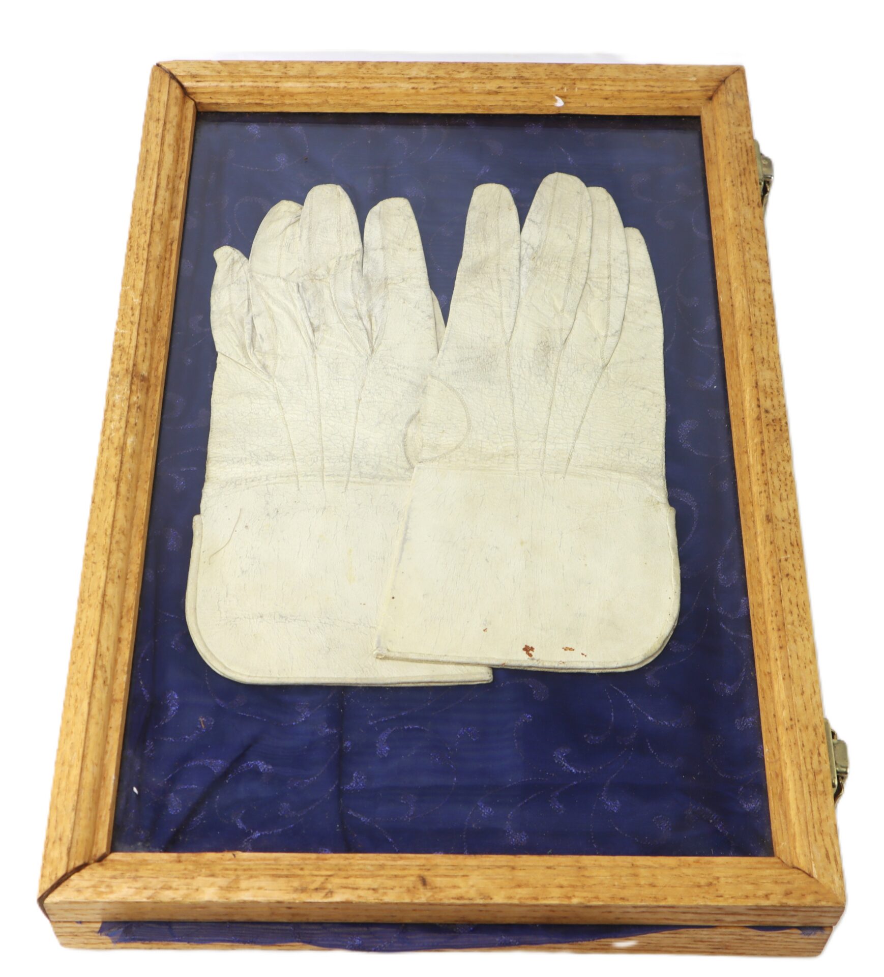 A pair of white gloves displayed in a wooden box with a glass top, resting on a blue fabric background.