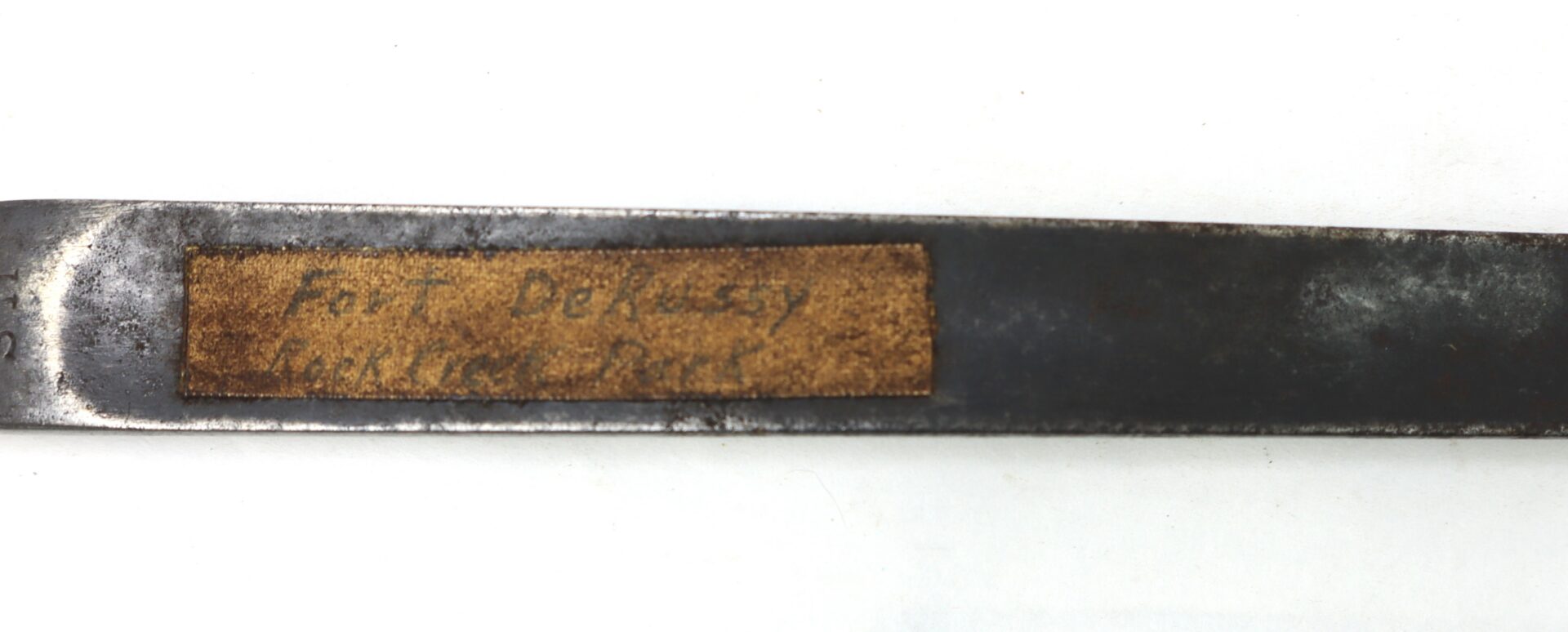 A close-up of a metal object with a worn, rectangular brown label that has faded handwritten text, partially reading "Fort Debussy" and "Honolulu, Hawaii.