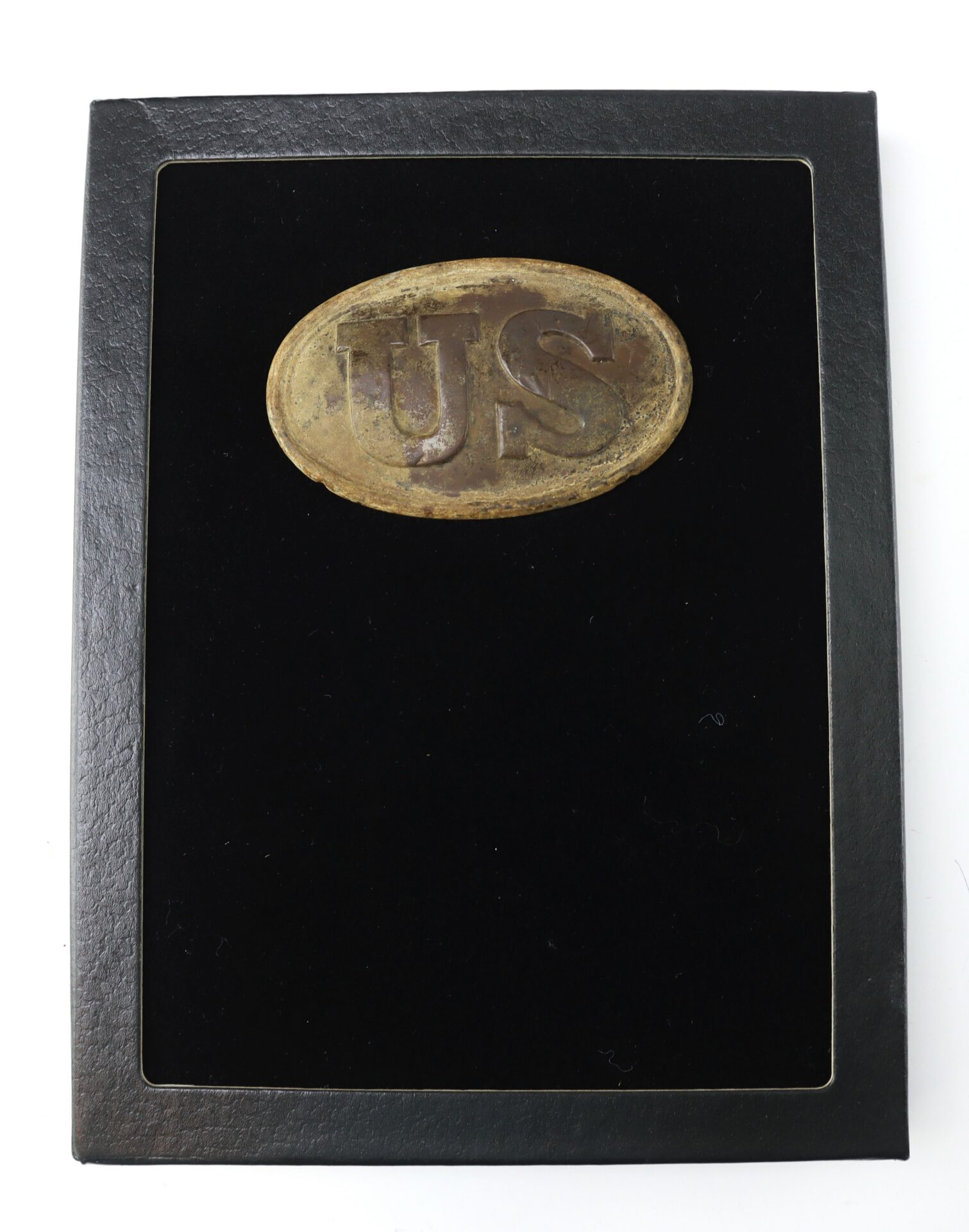An oval brass belt buckle marked "US" displayed on a black velvet background within a black frame.