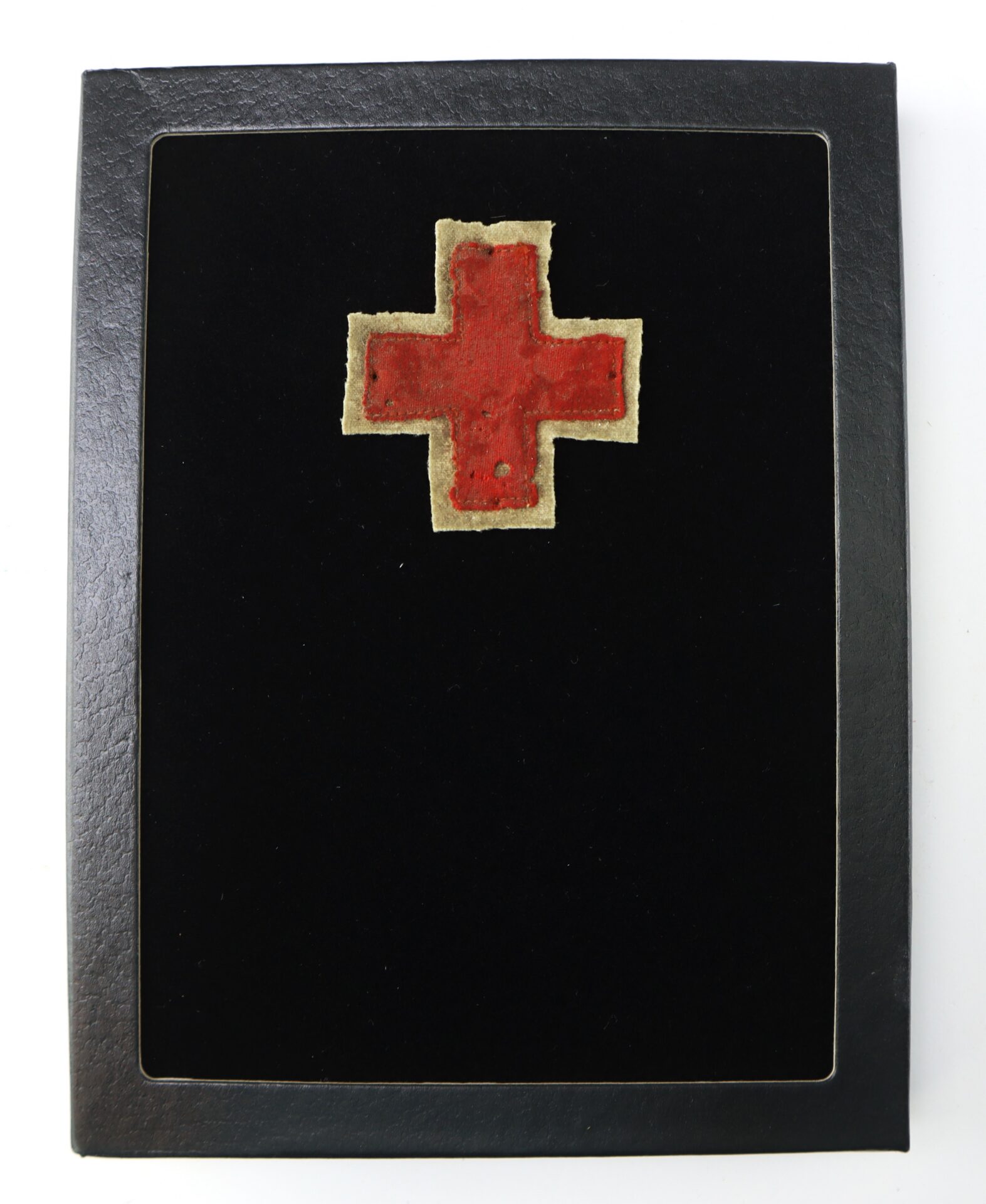 A red cross patch with a beige border is displayed on a black background inside a rectangular black frame.