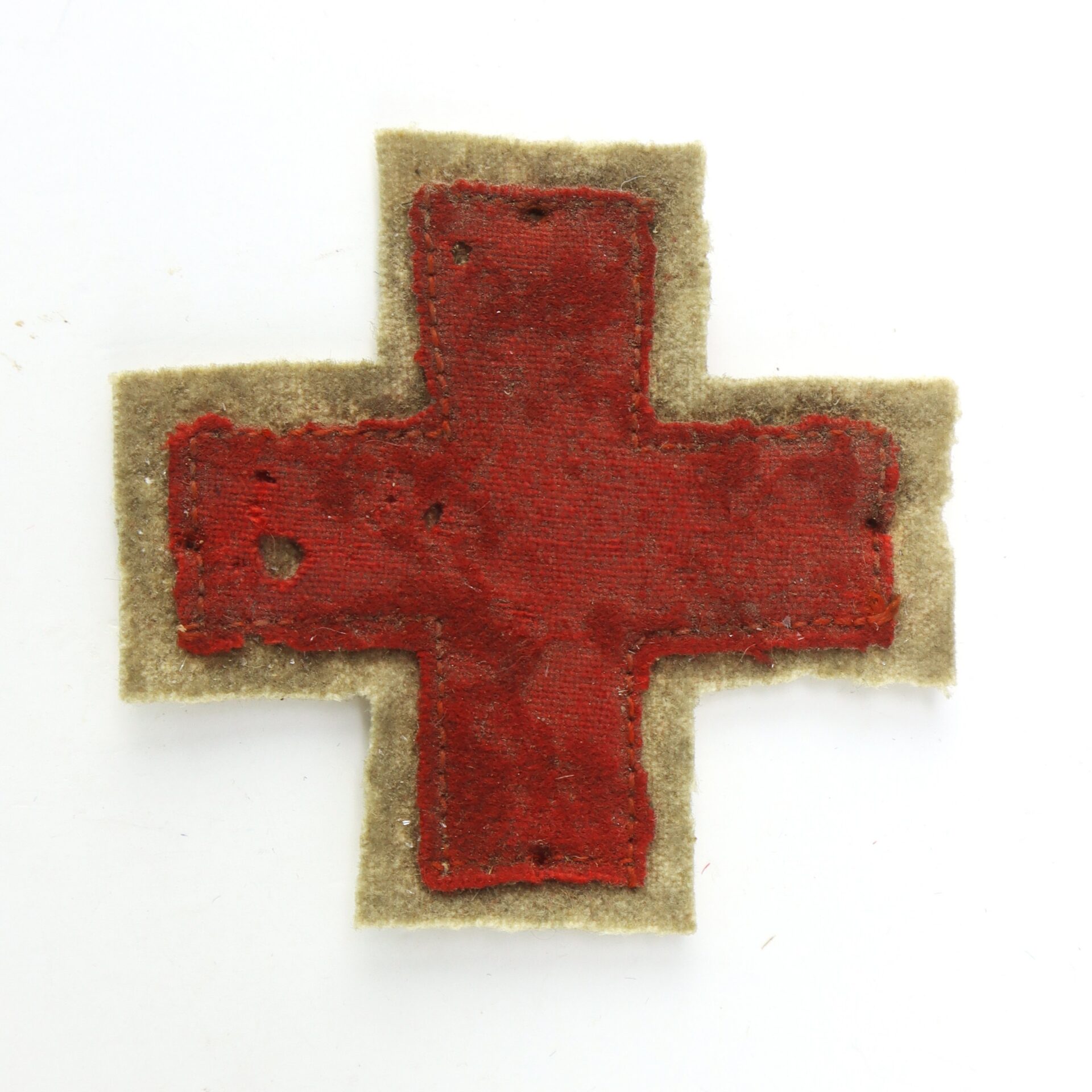 A fabric red cross patch with visible stitching on a beige background, showing signs of wear and minor fraying around the edges.