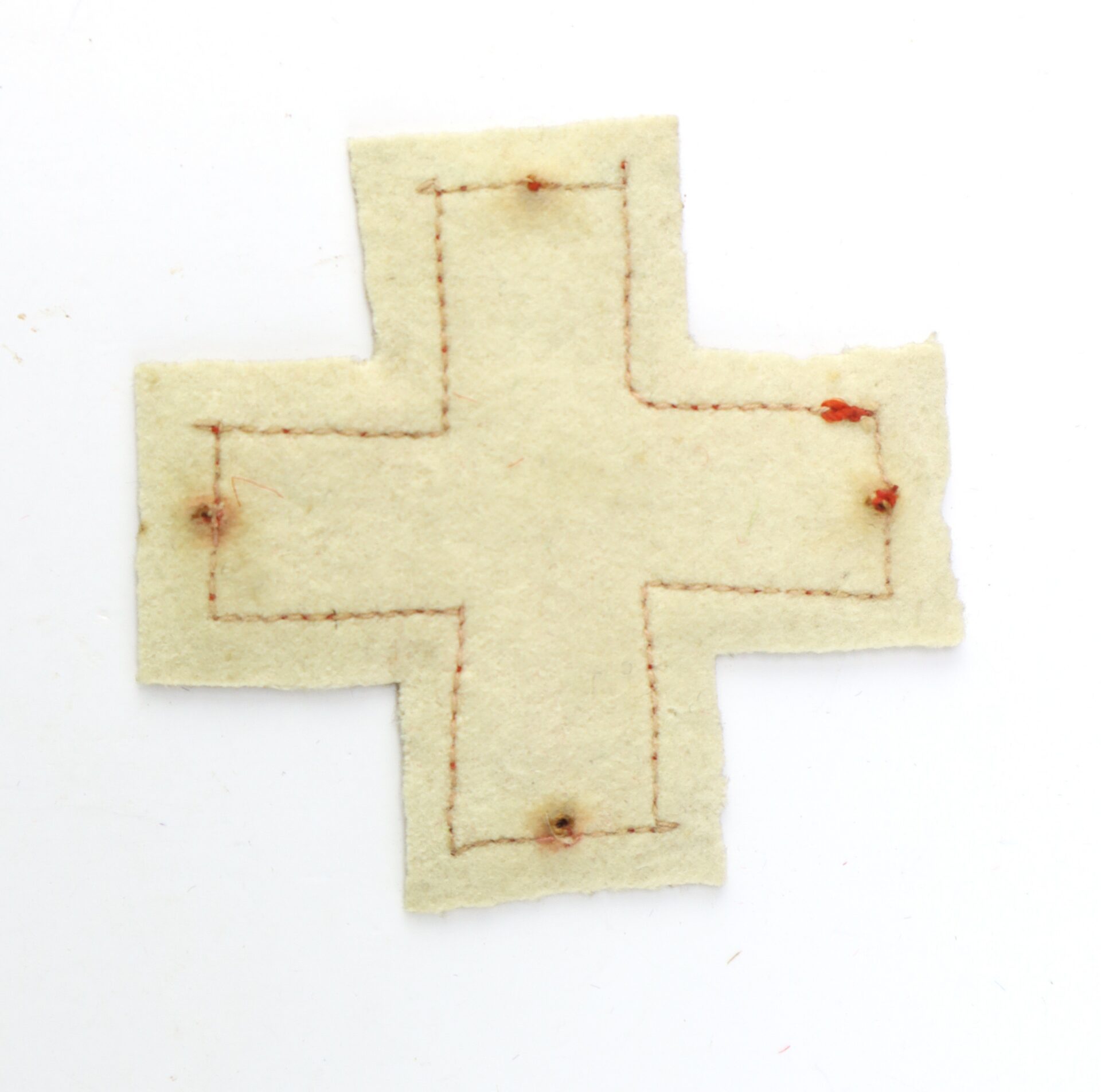 A piece of light-colored fabric cut into a cross shape with brown stitching and visible holes at several points.